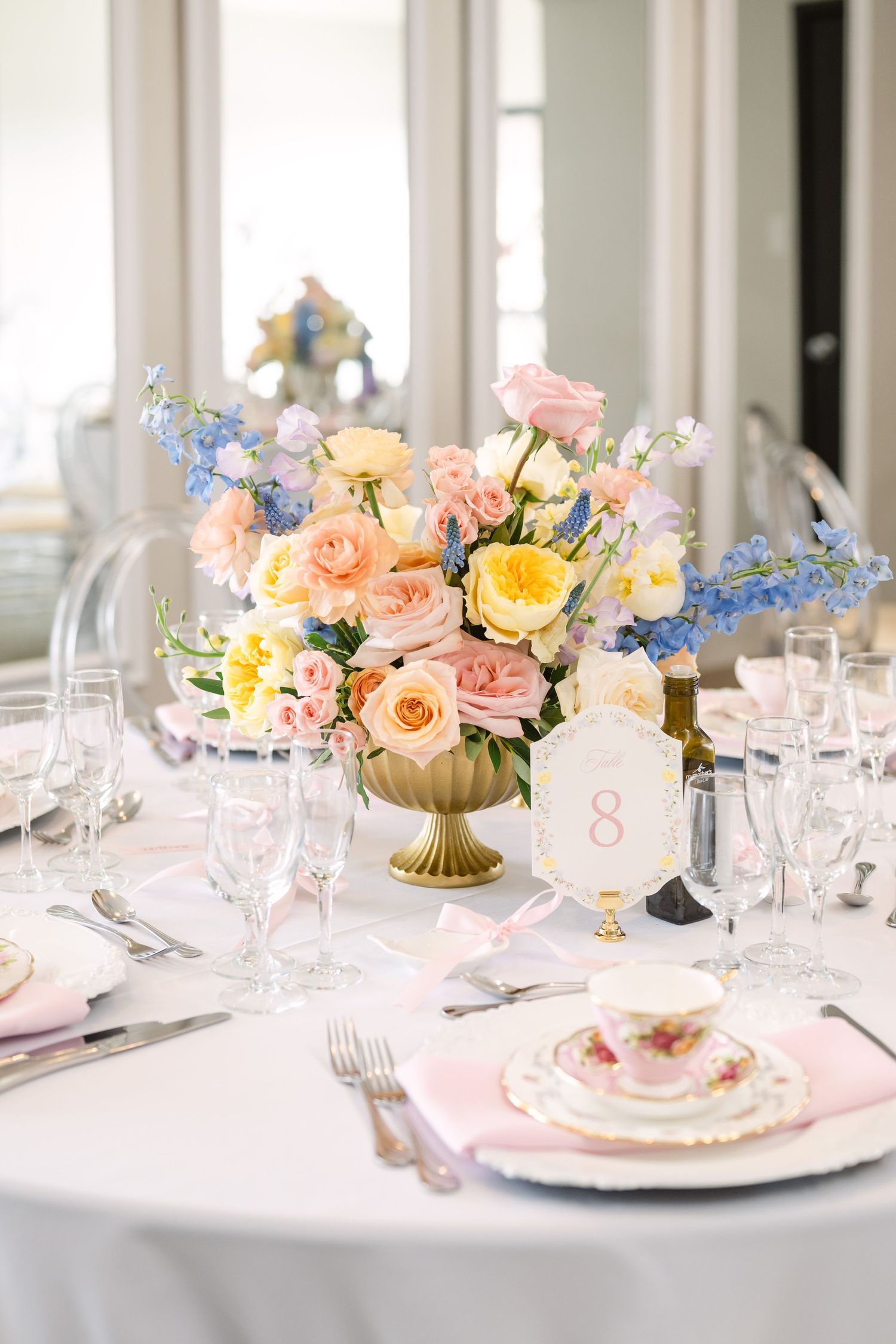 Elegant wedding reception table with pink, yellow, and blue floral centerpiece in gold vase, fine china, and crystal glassware.