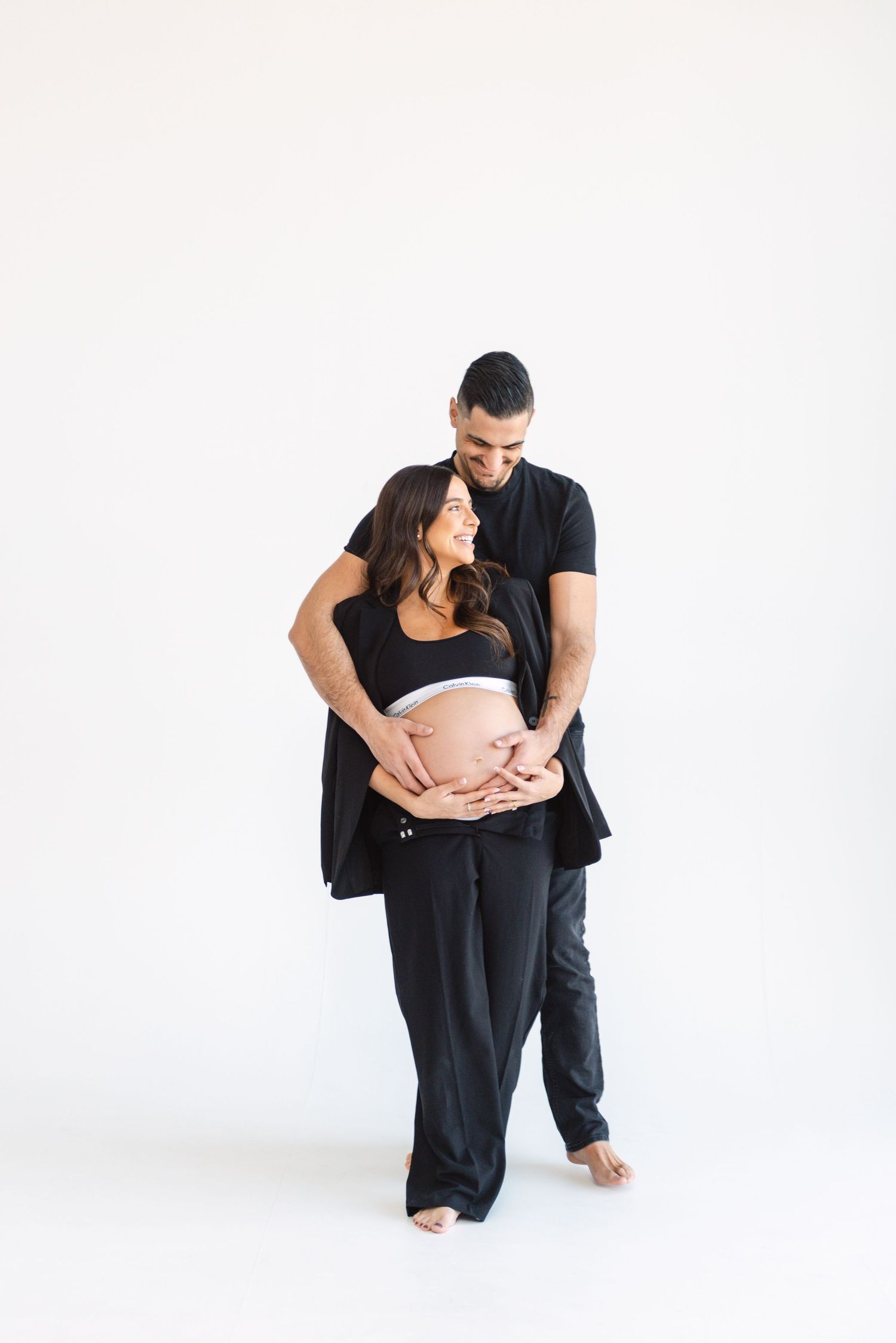 Intimate maternity photo of couple in black clothing embracing against white background.