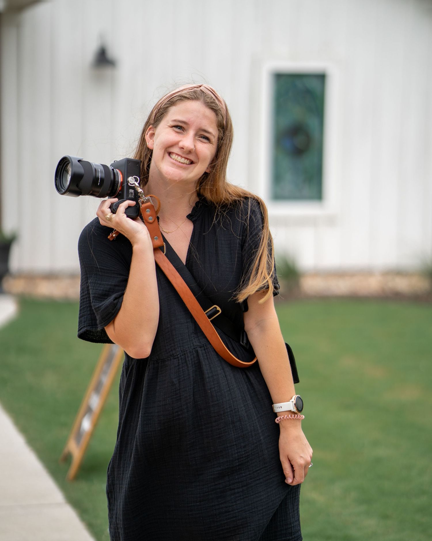 Professional photographer in black dress poses cheerfully with camera outdoors.