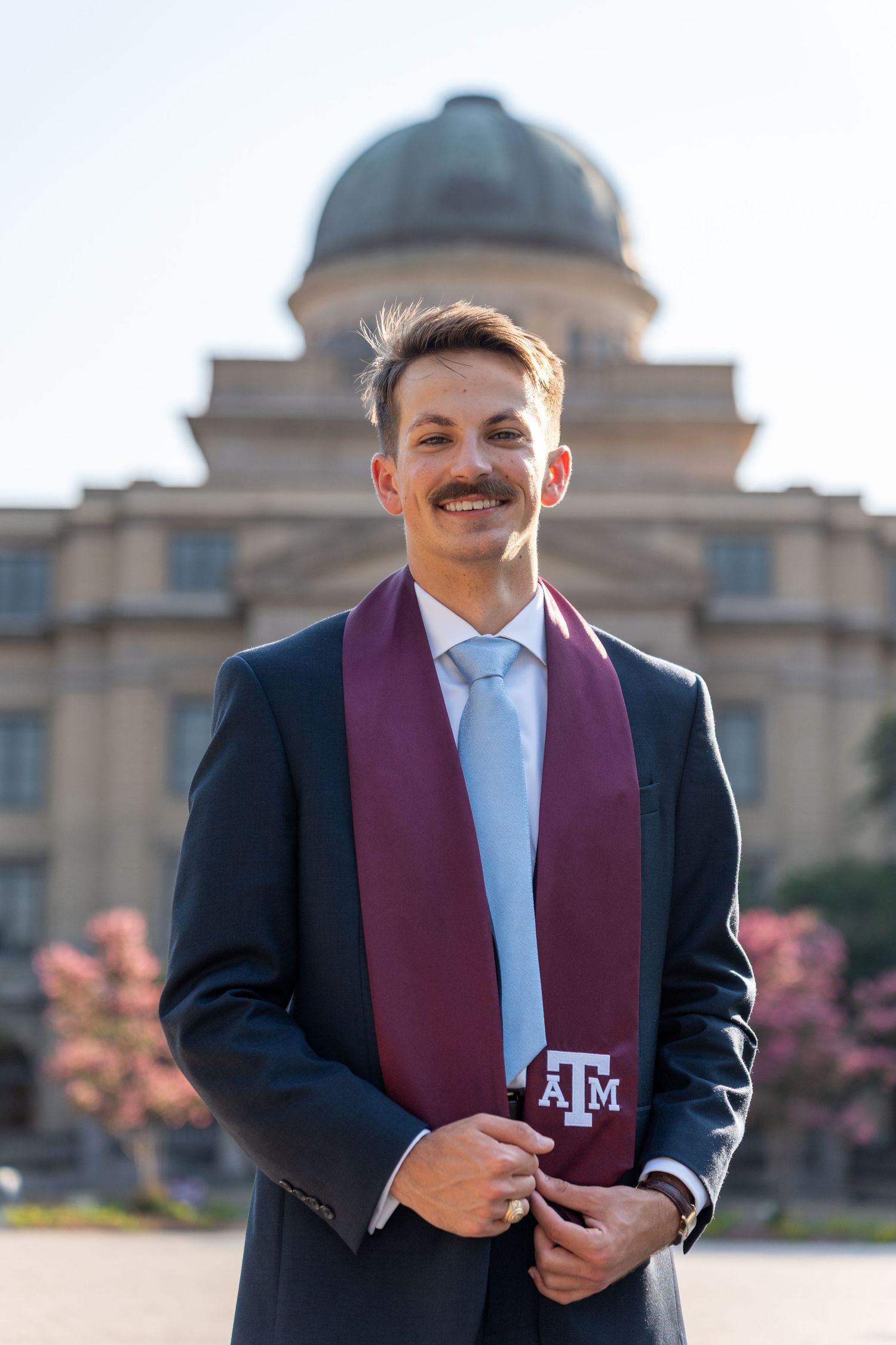 A graduate poses in a maroon Texas A&M stole and black cap against a collegiate building backdrop.