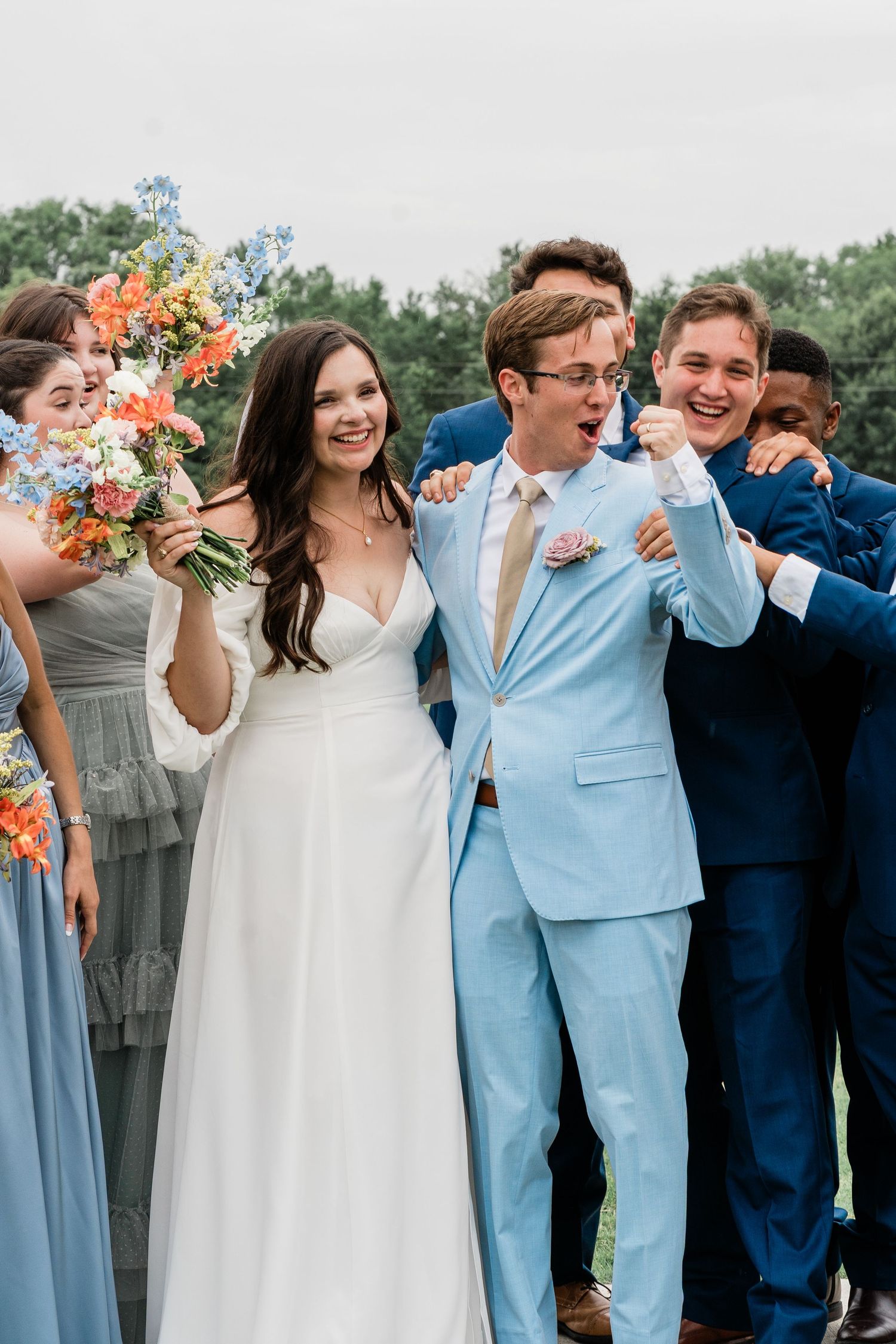 Wedding party celebrates outdoors with the groom in light blue and bridesmaids in sage green dresses.