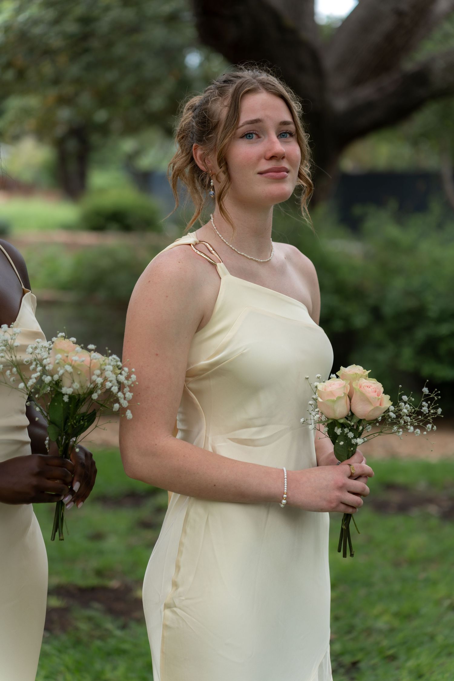 A bridesmaid in a pale yellow dress holds a white rose bouquet outdoors.