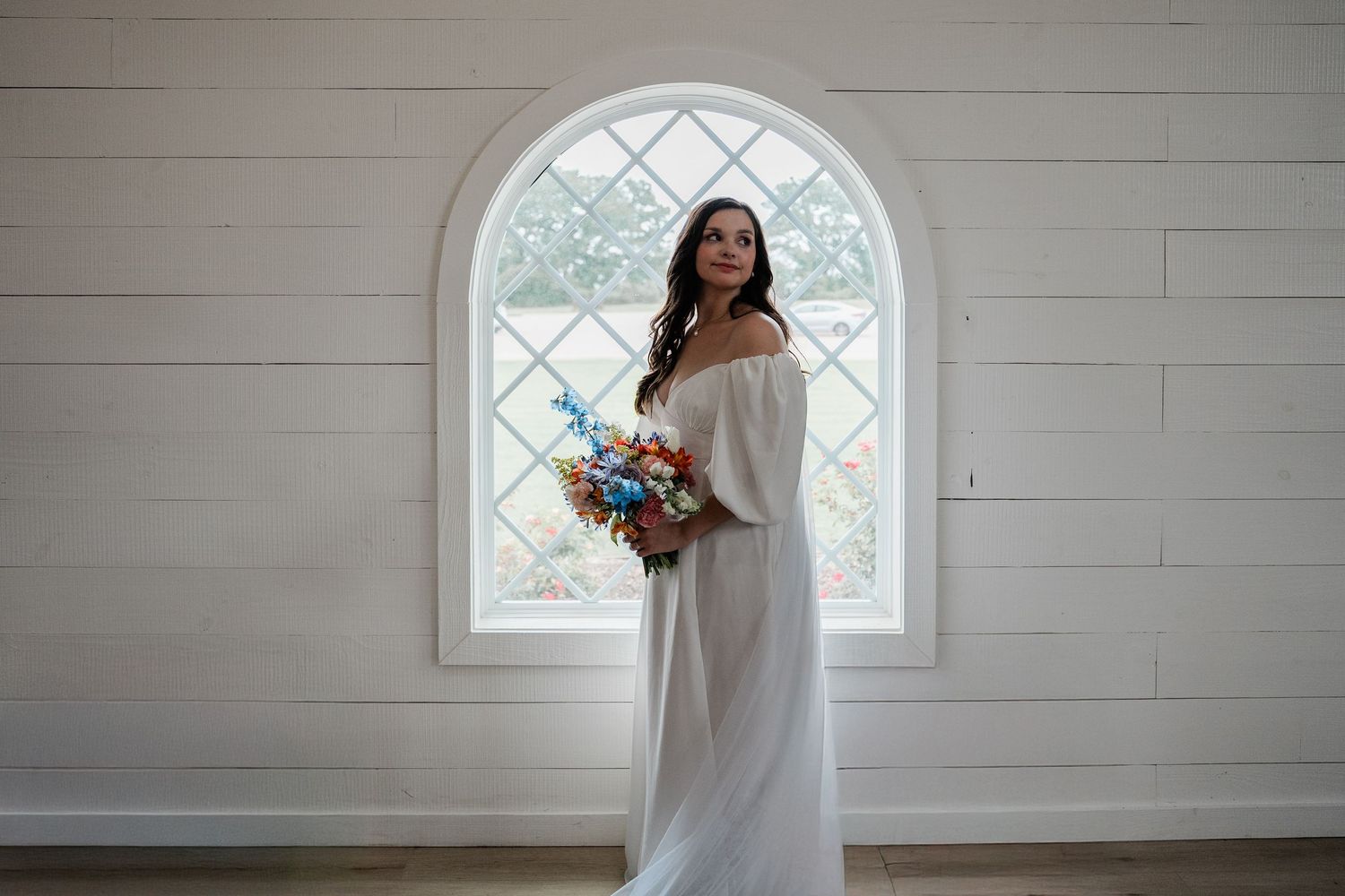 A white wedding dress with a floral bouquet featuring blue and orange flowers against a light interior wall.