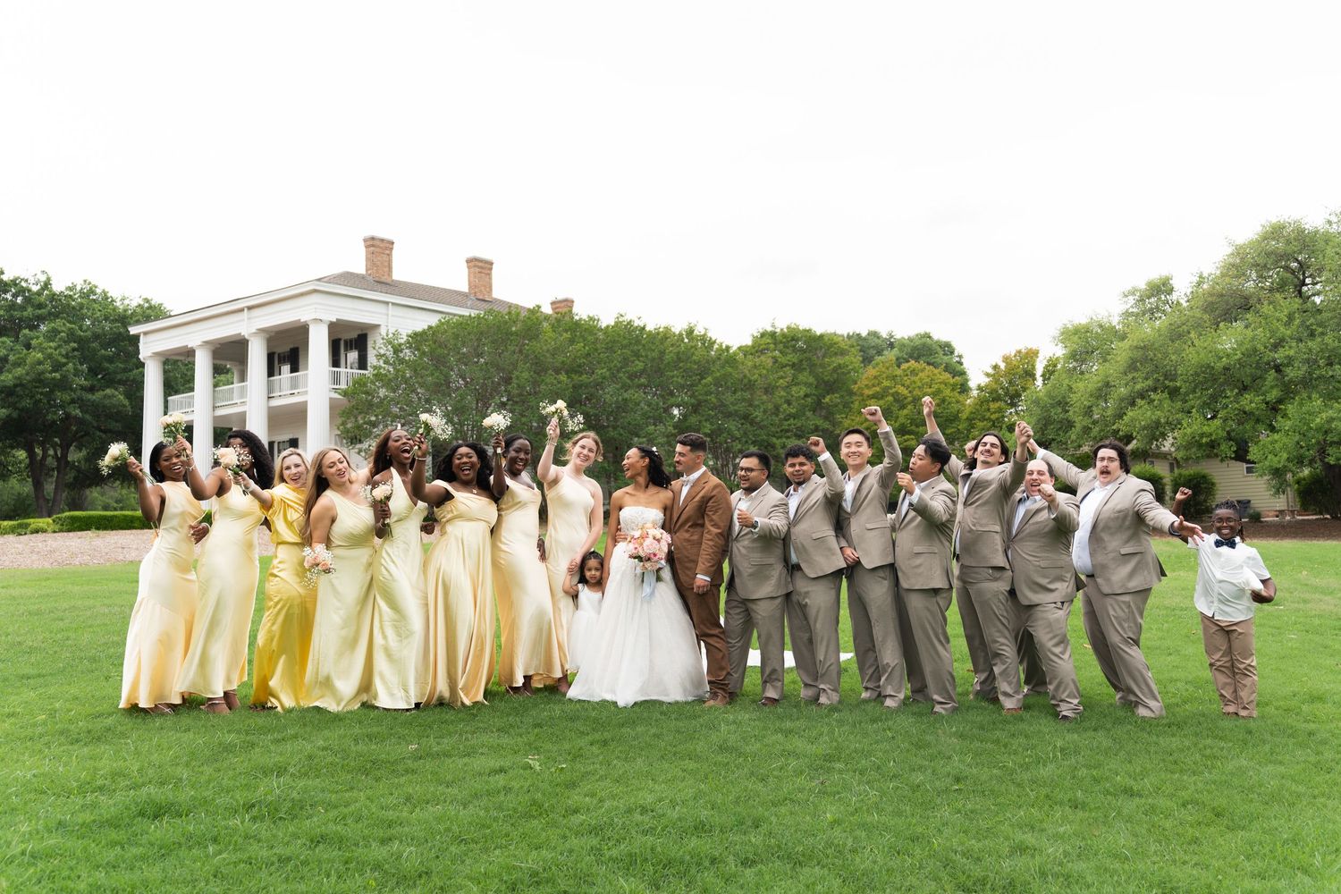 Wedding party in yellow dresses and gray suits posing on a green lawn with white mansion in background.