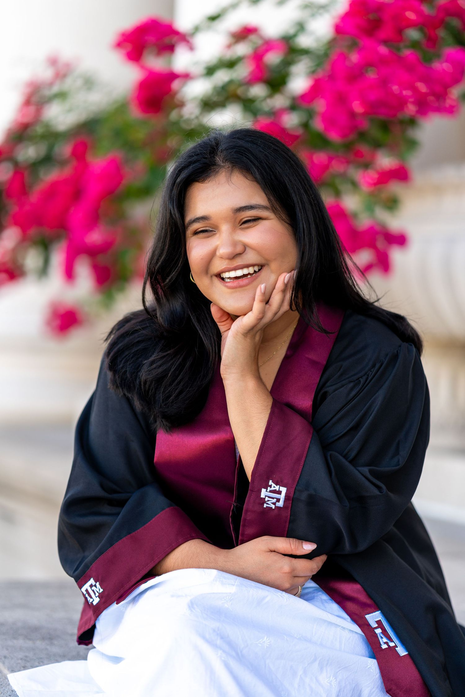 A joyful graduate in black and maroon regalia poses with bright pink bougainvillea flowers in the background.