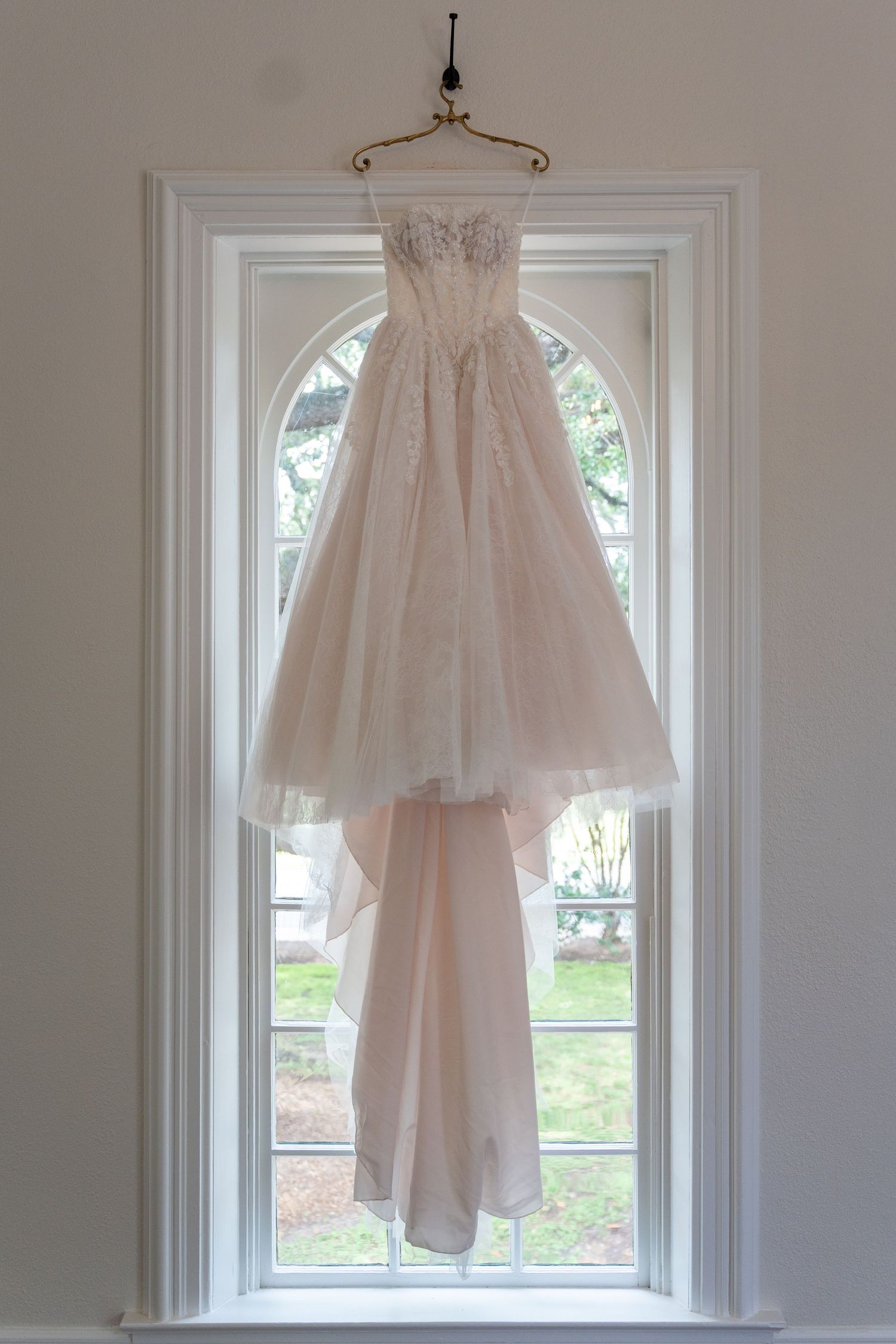 A delicate blush wedding dress hanging in front of a bright window with white trim.