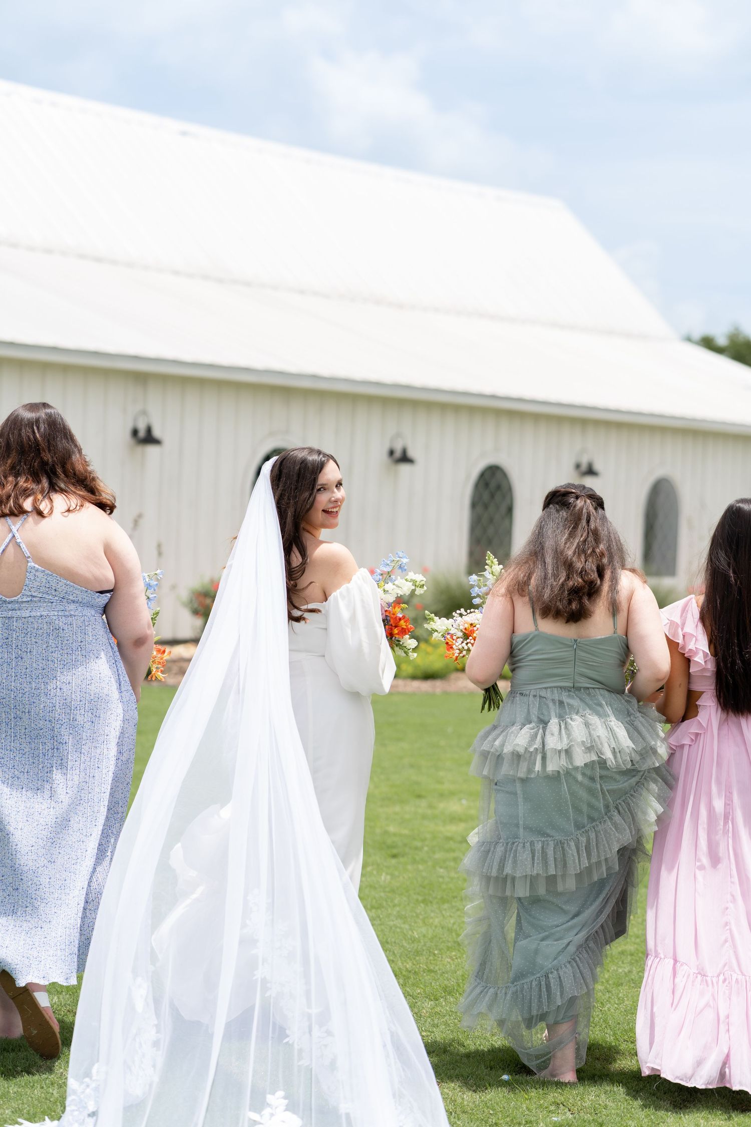 Wedding party members in pastel dresses gather outside a white barn venue.