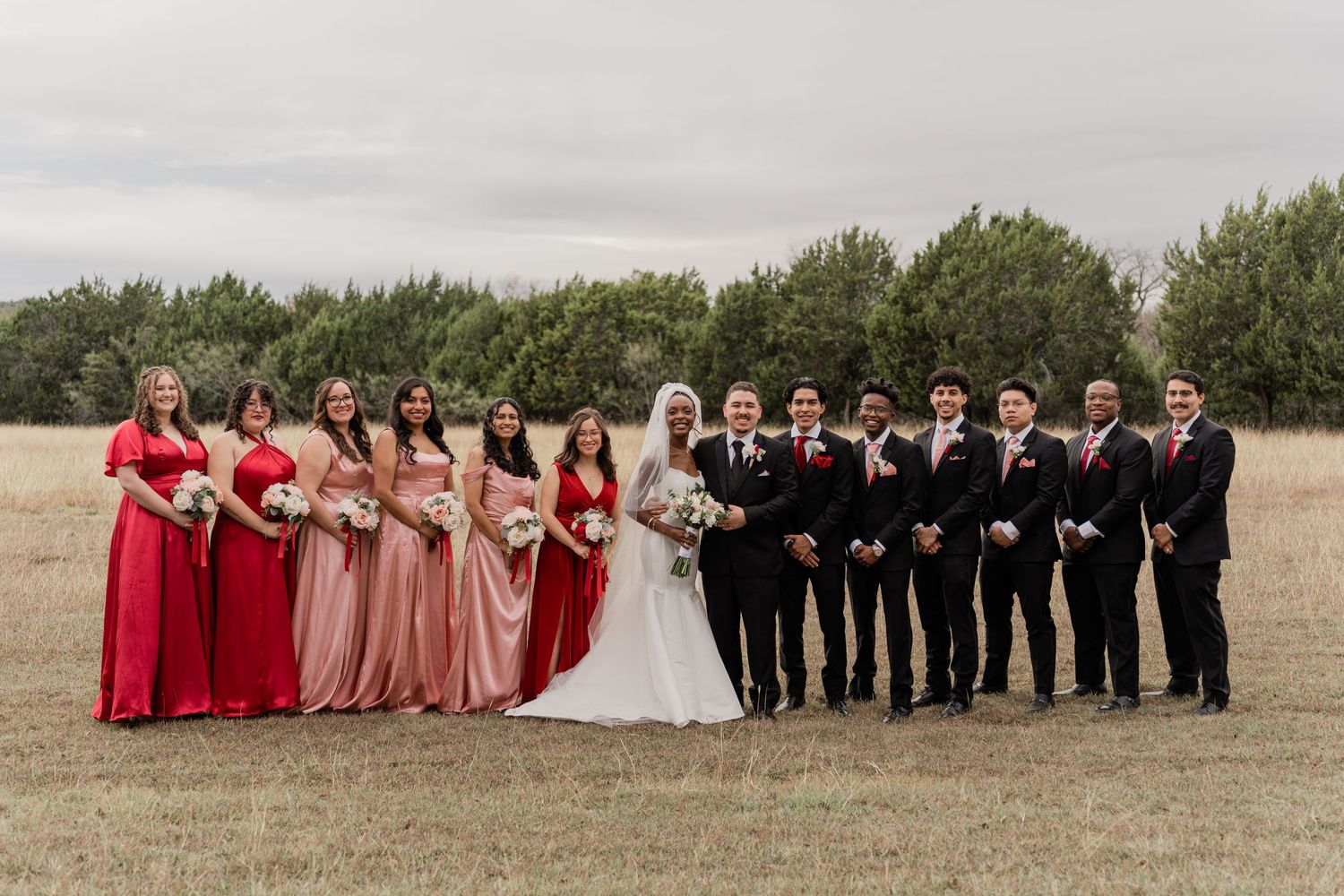 Wedding party in red and blush dresses poses with groomsmen in black suits on a grassy field during sunset.