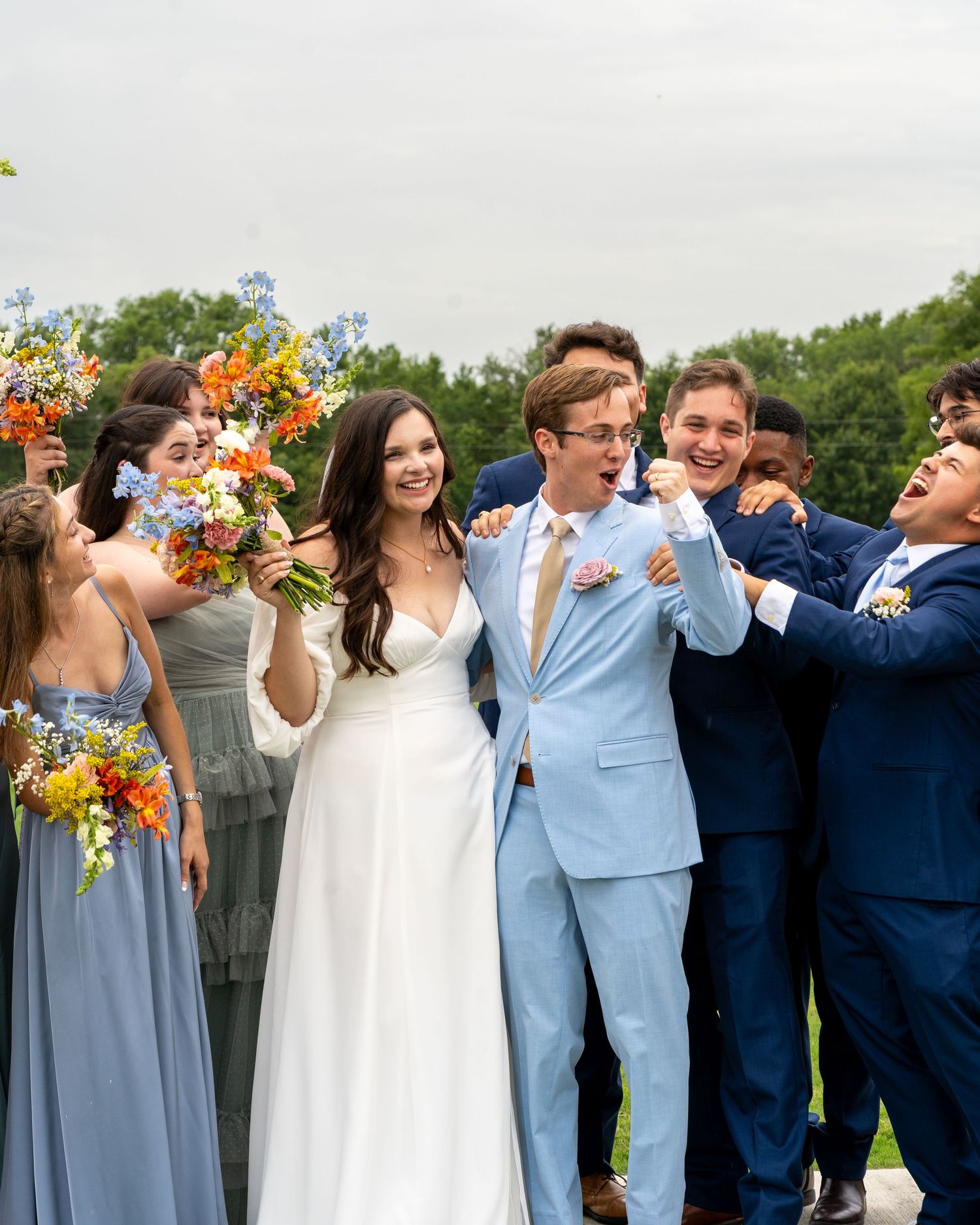 Wedding party celebrating with colorful sunflower bouquets and blue bridesmaid dresses outdoors.