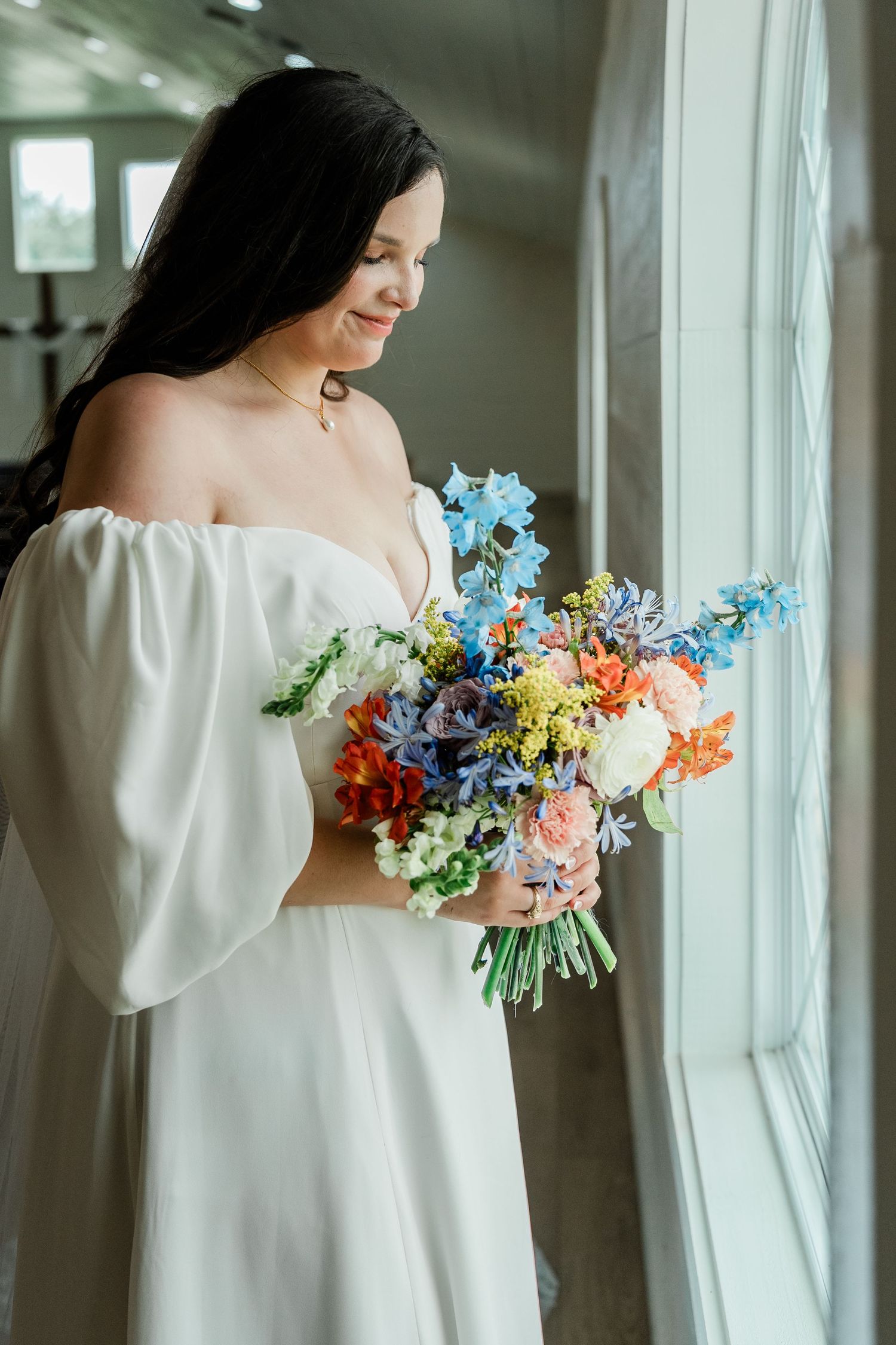An elegant white wedding dress with flowing sleeves and a vibrant mixed floral bouquet by a window.