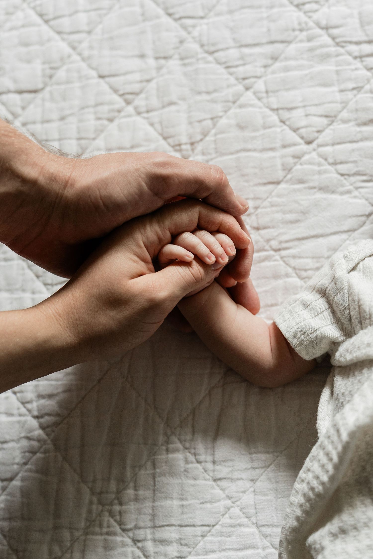 Tender close-up of parent holding newborn baby's hand on white quilted blanket.