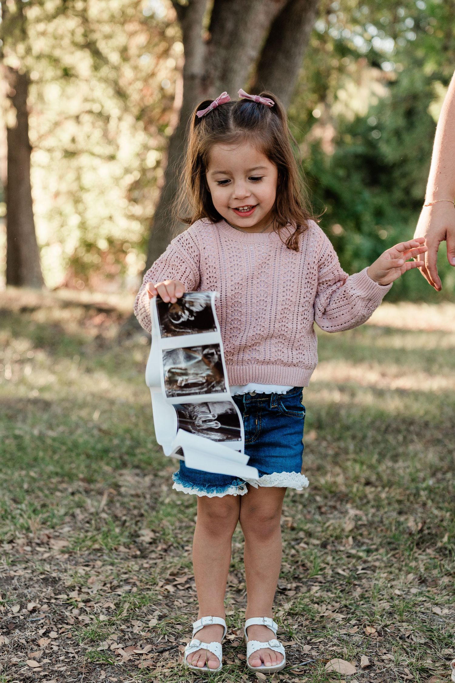 Young child in pink sweater holding ultrasound photos while standing on autumn leaves.