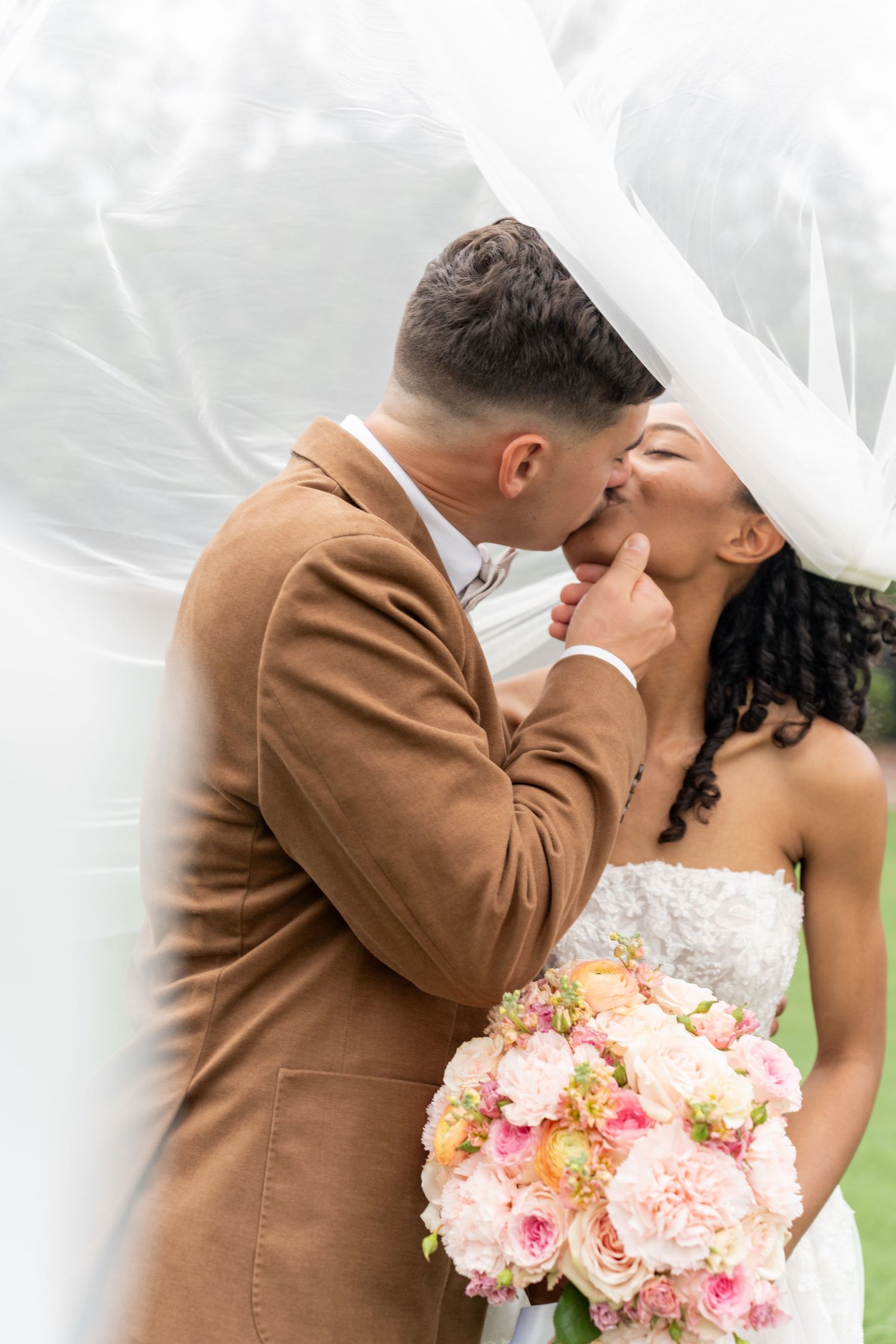 Romantic wedding couple shares intimate kisses while bride holds pink and peach floral bouquet.