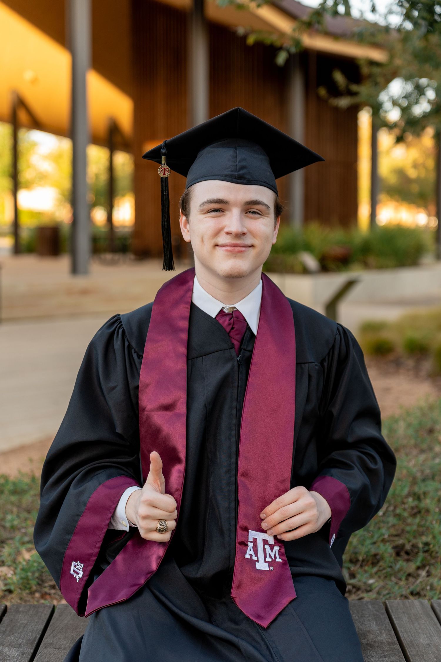 Graduate in black cap and gown with maroon stole smiles during outdoor graduation photo session.