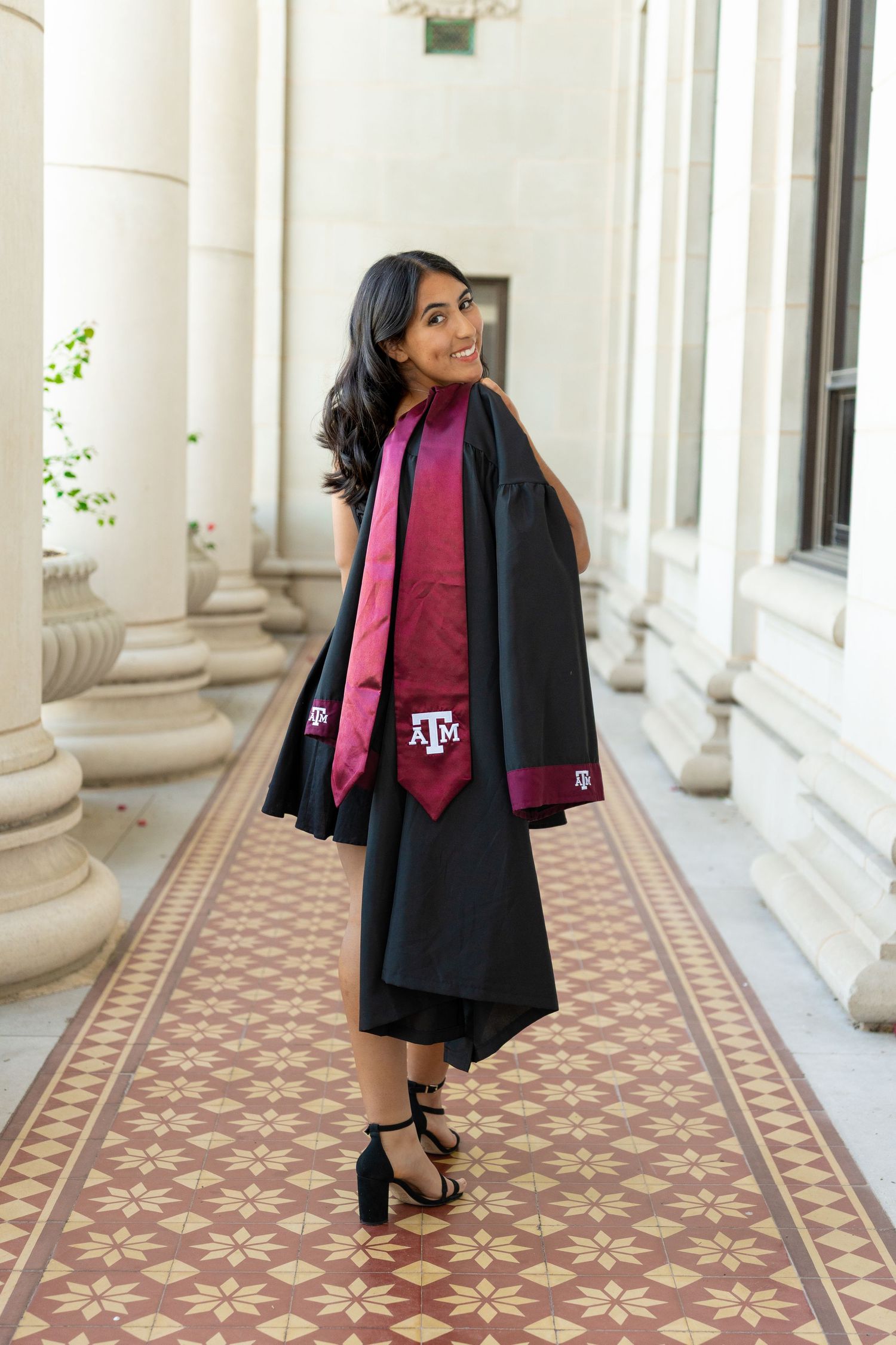 Graduate in black gown and maroon tie poses elegantly in columned hallway.