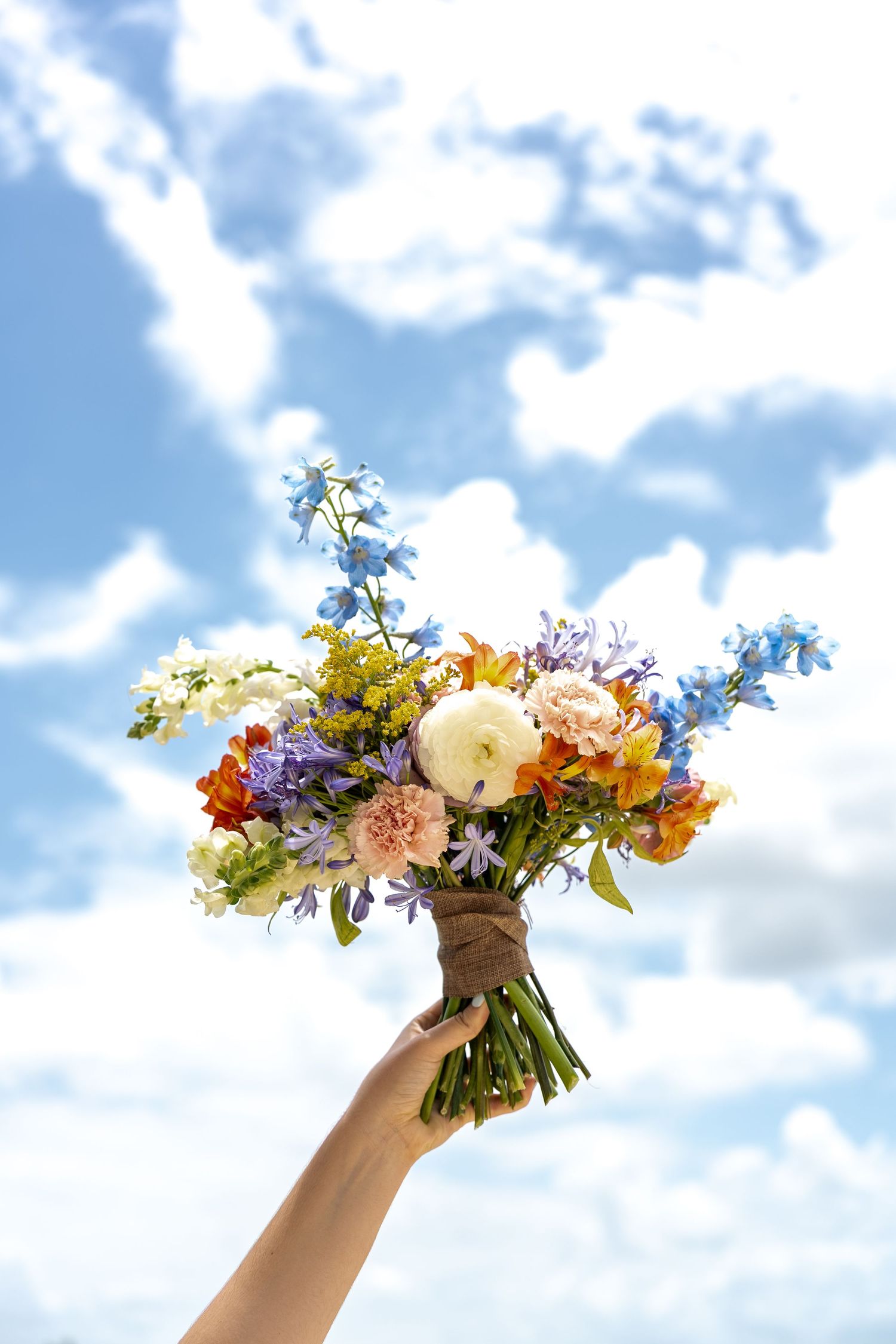 Hands holding colorful wildflower bouquets against a blue sky with white fluffy clouds.