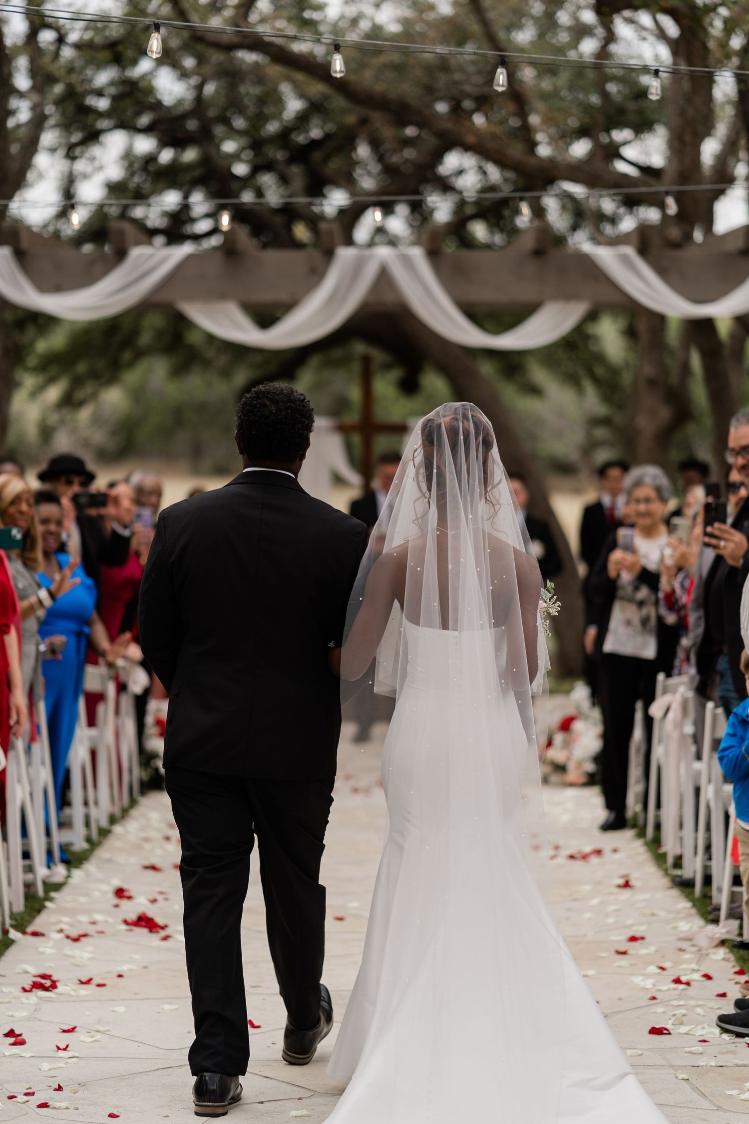 Bride and groom walk down outdoor wedding aisle lined with rose petals and white chairs as guests look on.