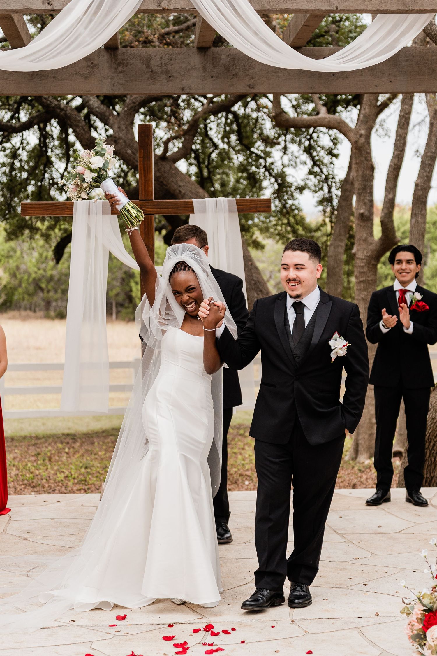 A wedding party celebrates and dances together under white fabric draping at an outdoor venue decorated with red rose petals.