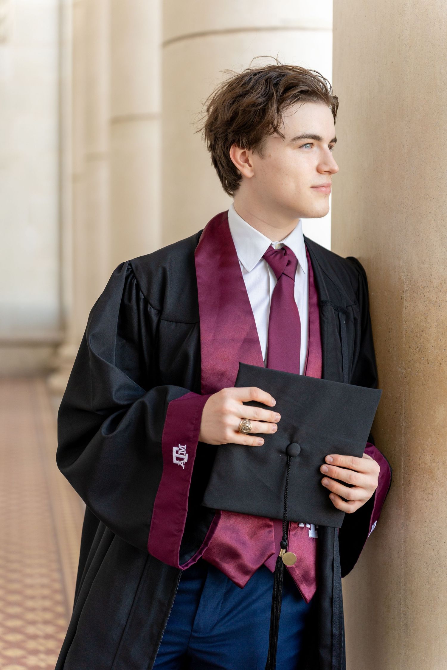 Graduate in black academic gown and burgundy stole holds diploma while looking away in classic portrait pose.