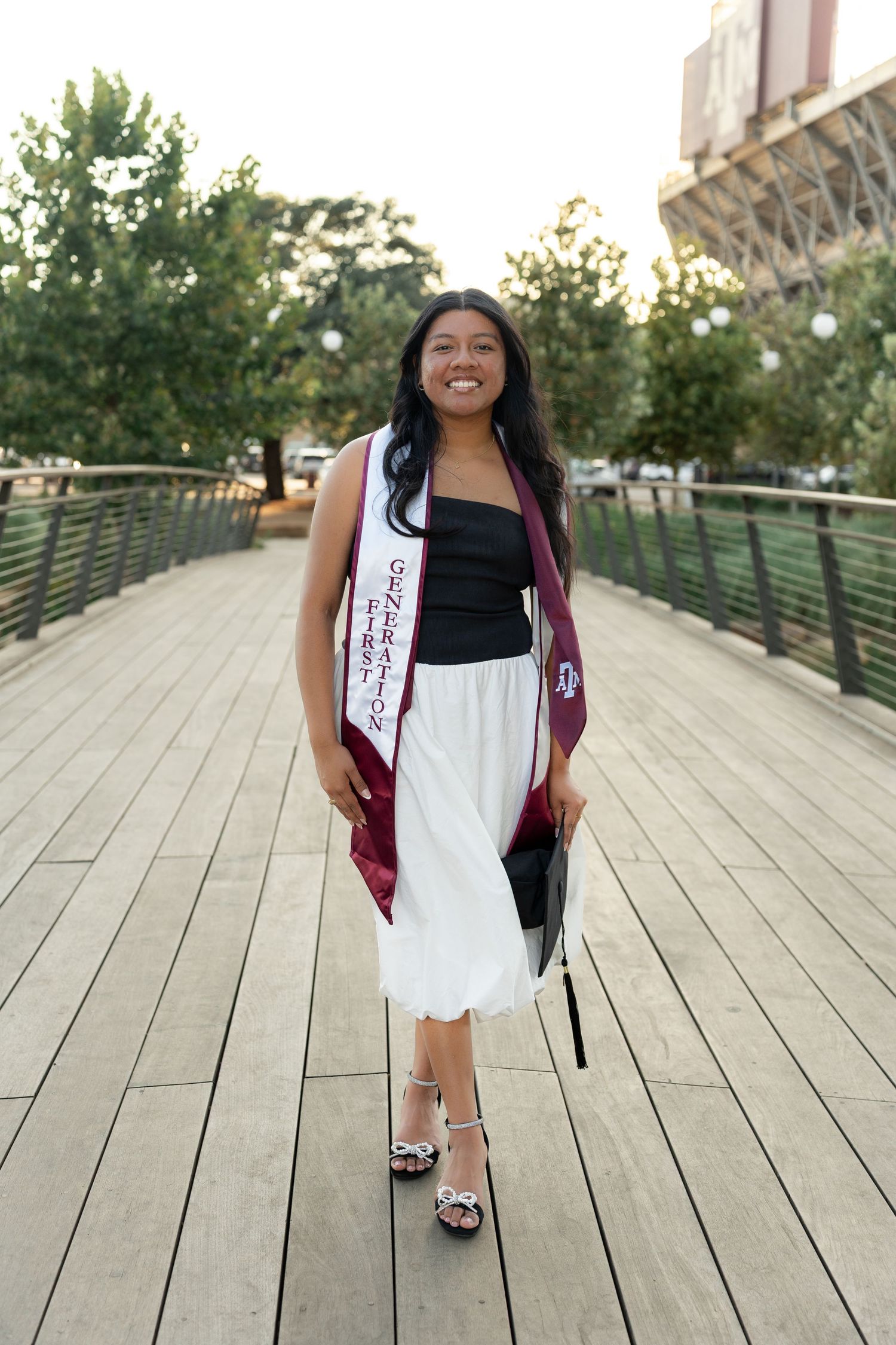 A person in a burgundy cardigan and white dress poses on a wooden deck walkway with a bright smile.