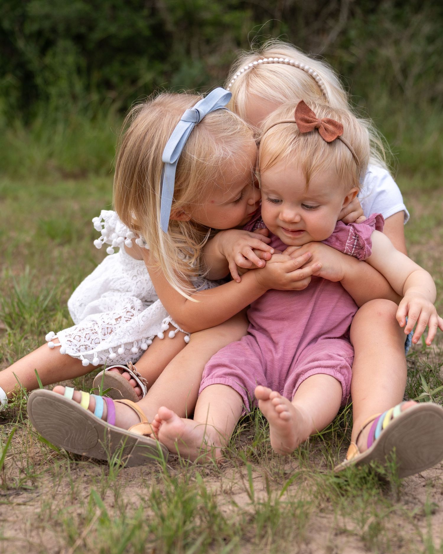 Two young children in light colored outfits share sweet moments while sitting together outdoors in nature.