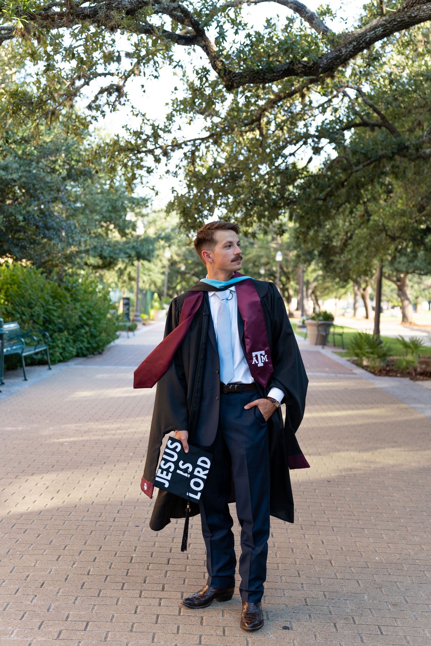 A graduate in academic regalia walks along a brick pathway under oak trees on a sunny day.