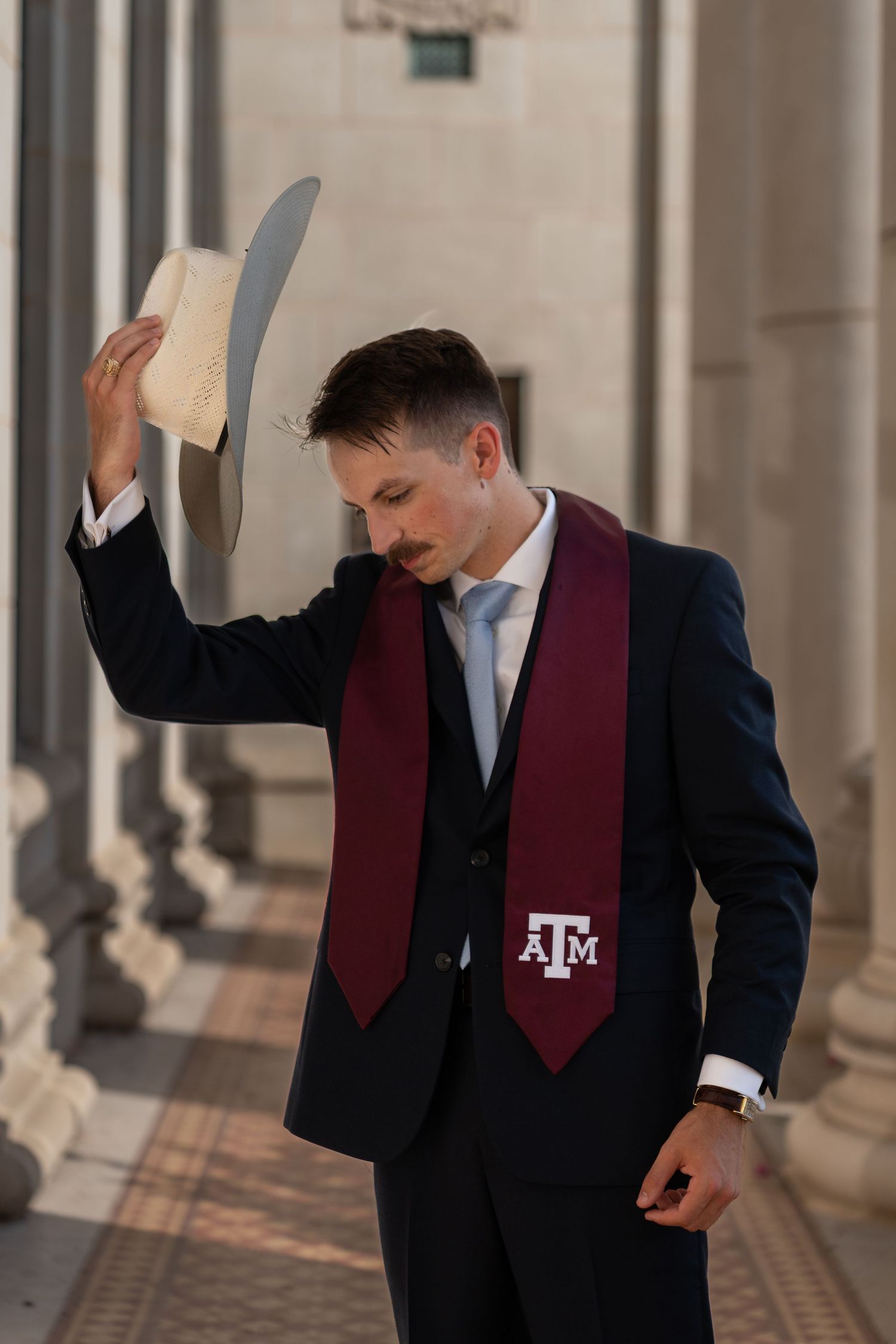 Graduate wearing navy suit and maroon stole poses against architectural columns in celebratory graduation photos.