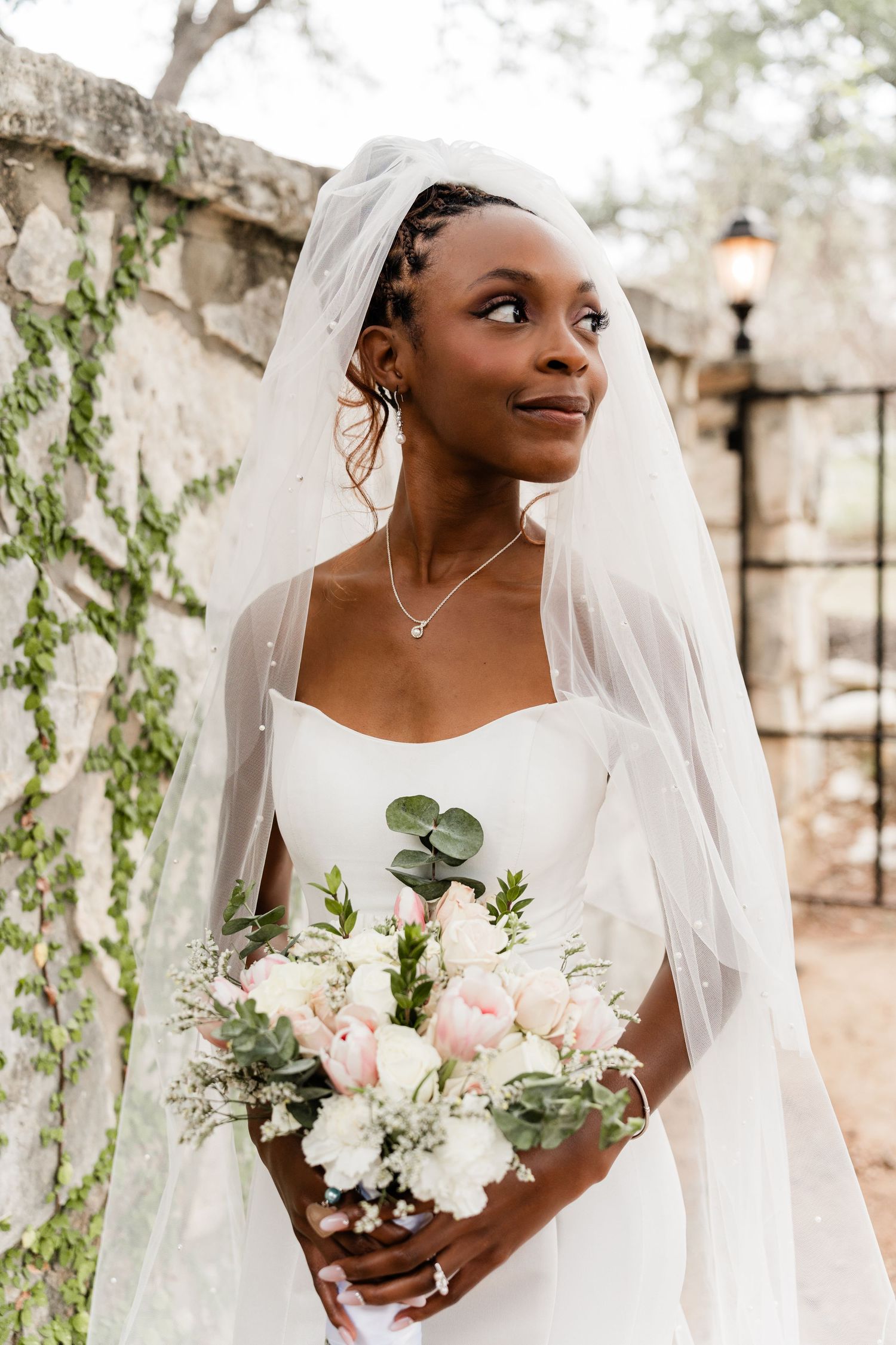 Beautiful bride in white wedding dress holding delicate bouquet of white and pink flowers with eucalyptus against stone wall.
