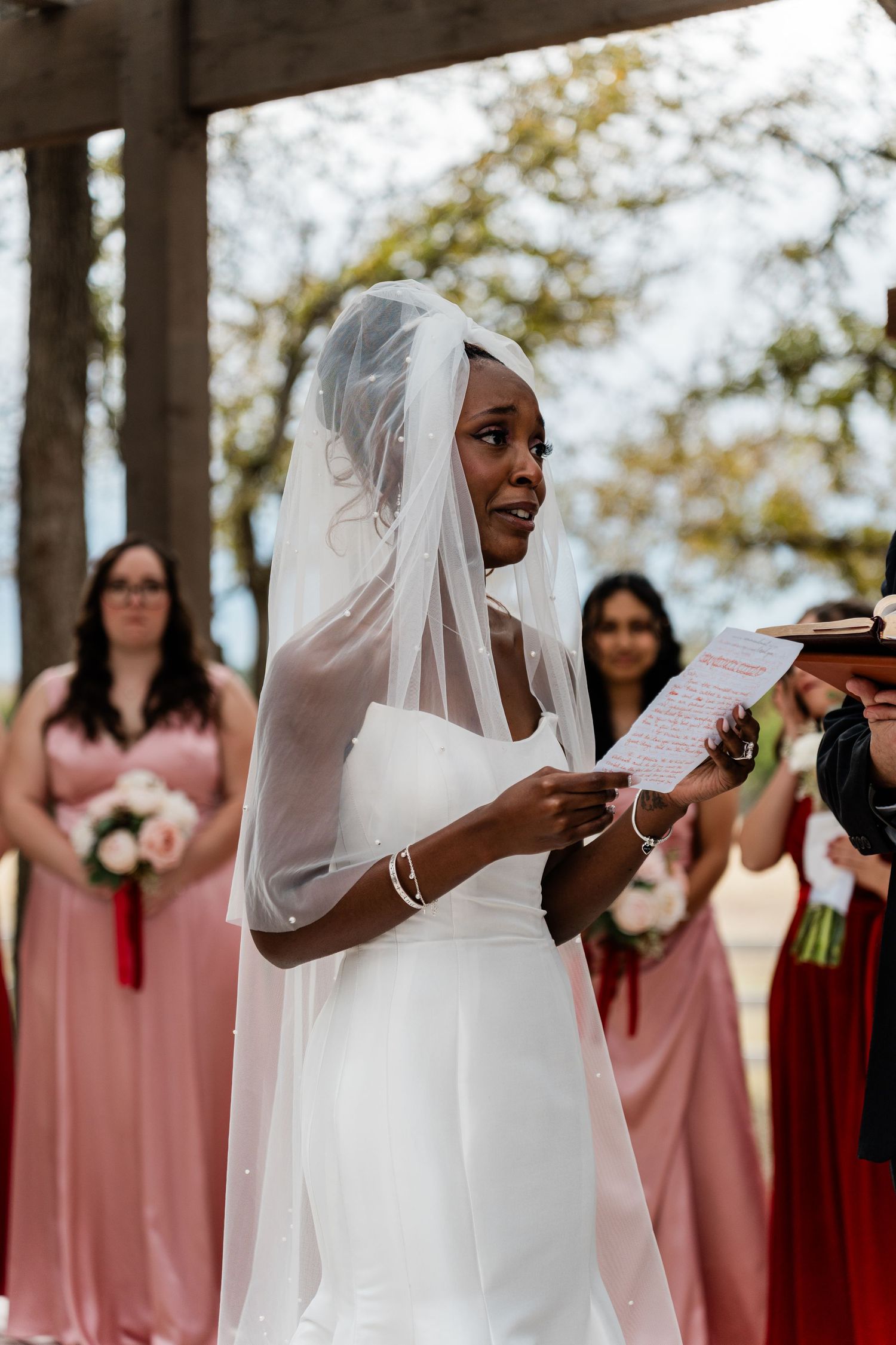 Bride in white dress stands with bridesmaids wearing pink dresses during an outdoor wedding ceremony.