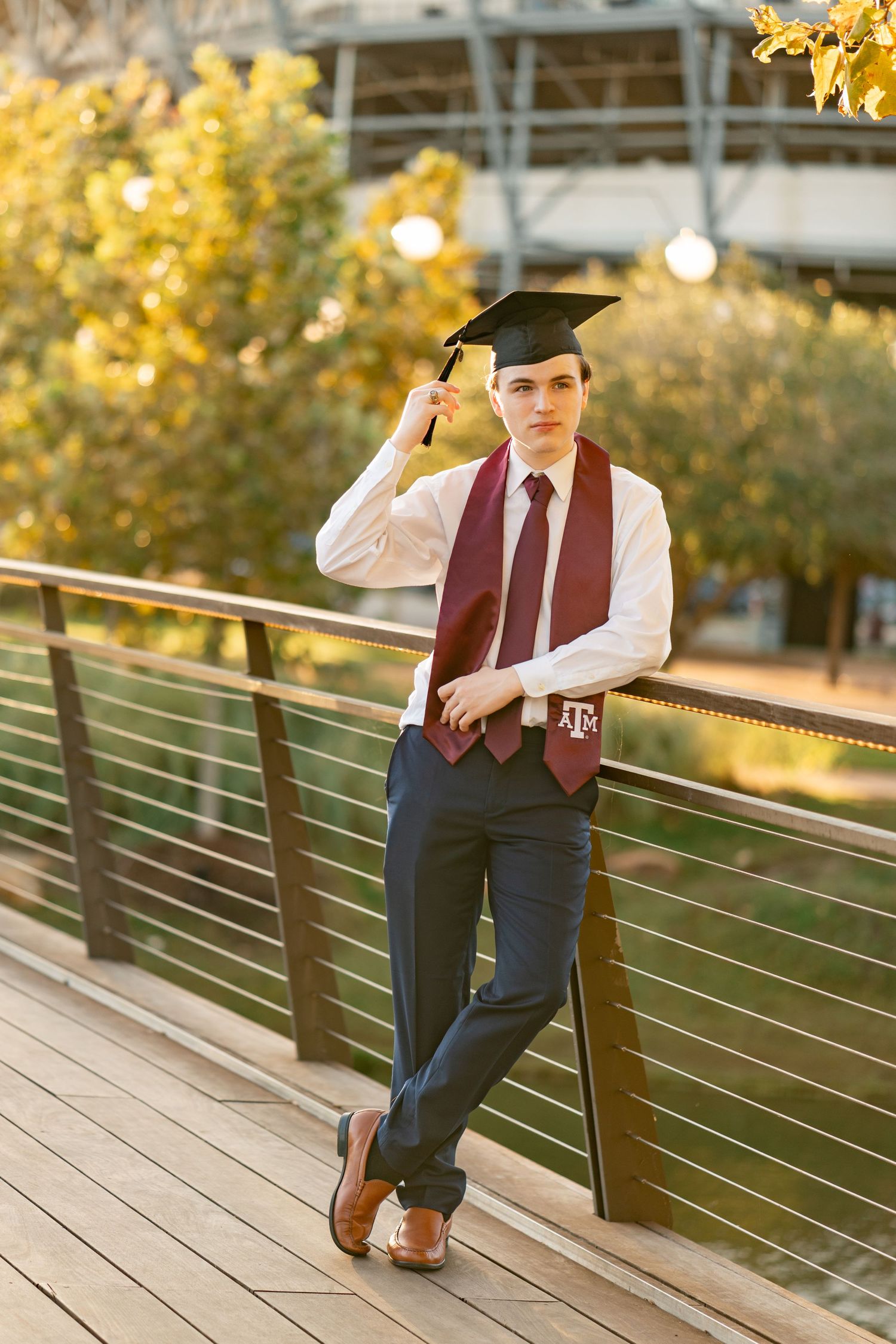 A graduate in a maroon vest and graduation cap poses on a wooden deck during golden hour.