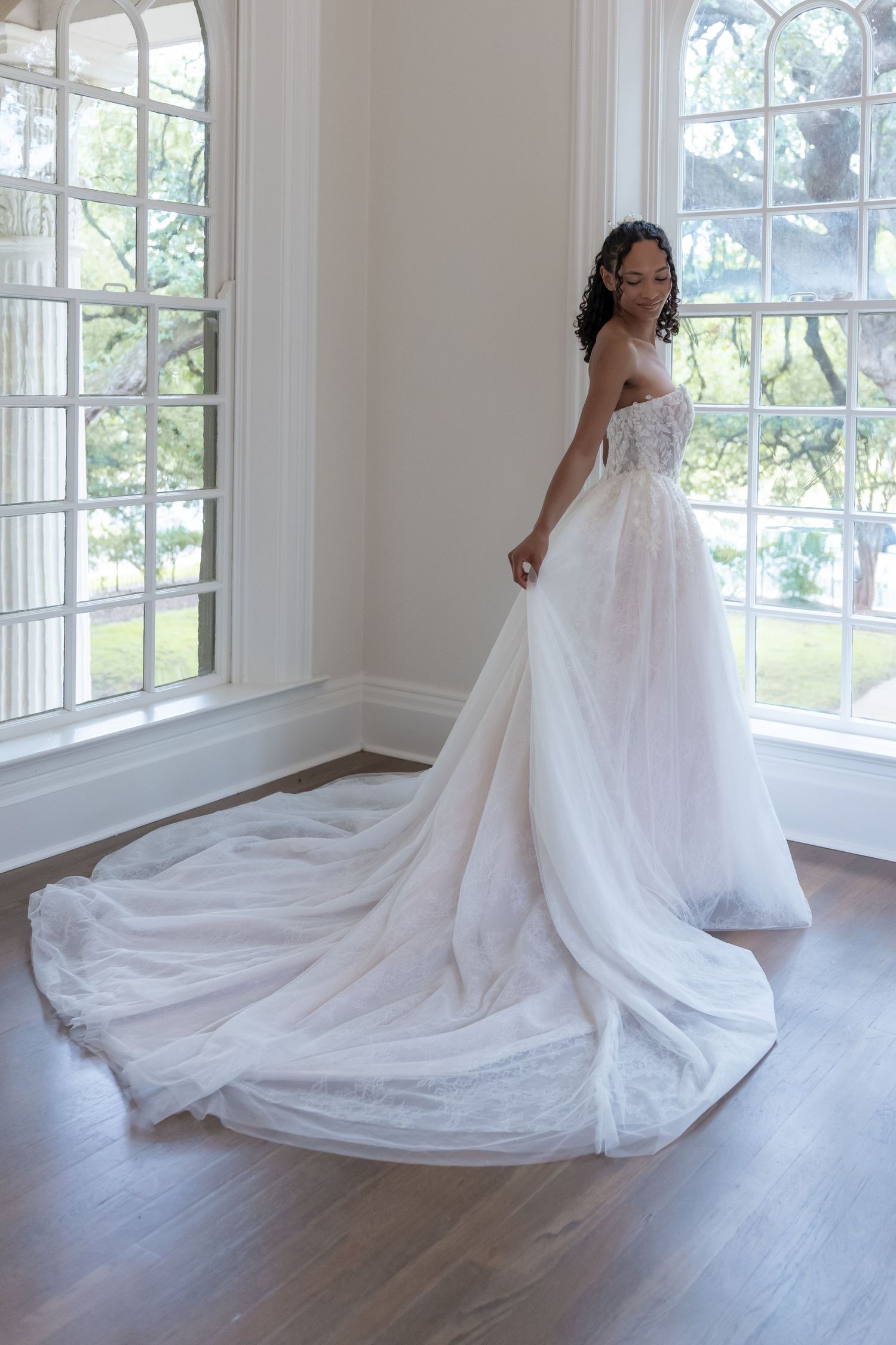 A flowing white wedding dress with a long train displayed in a bright sunlit room with hardwood floors.