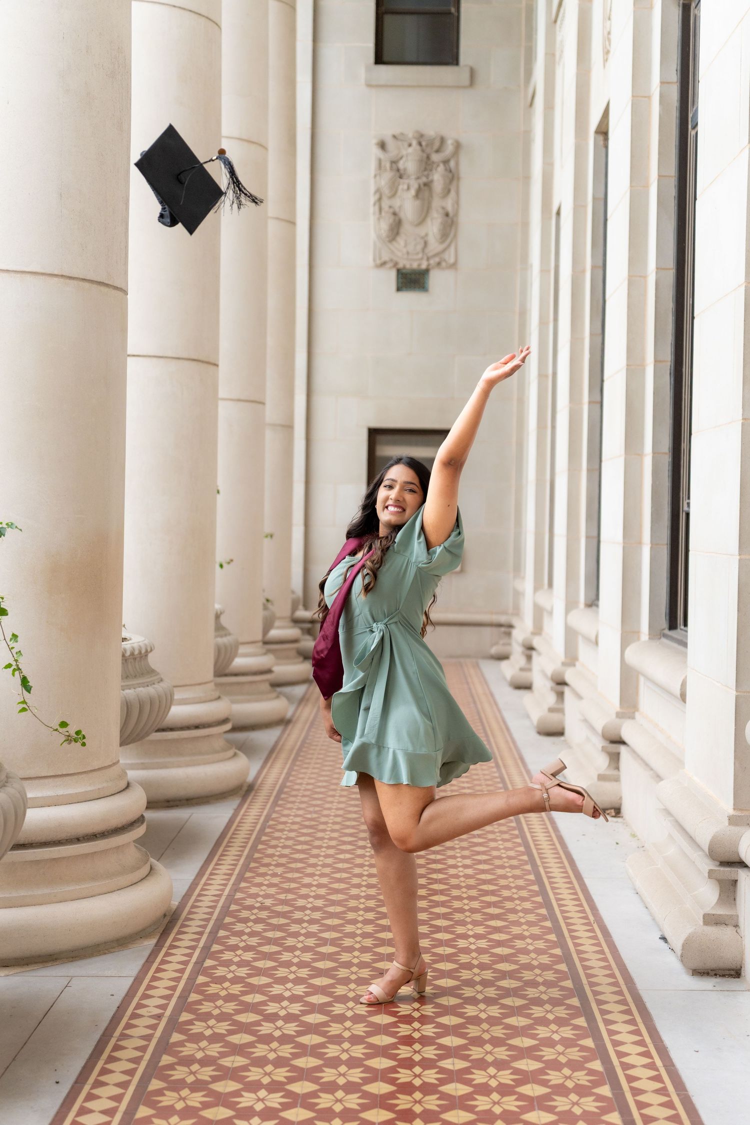 Graduate in mint green dress throwing cap in ornate hallway with classical columns.