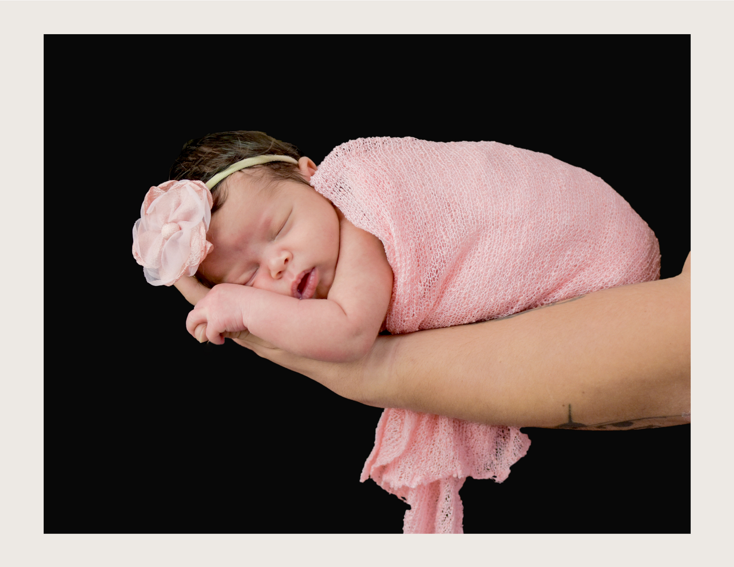 Newborn baby girl sleeping peacefully in pink wrap and floral headband, cradled in an adult's hands.