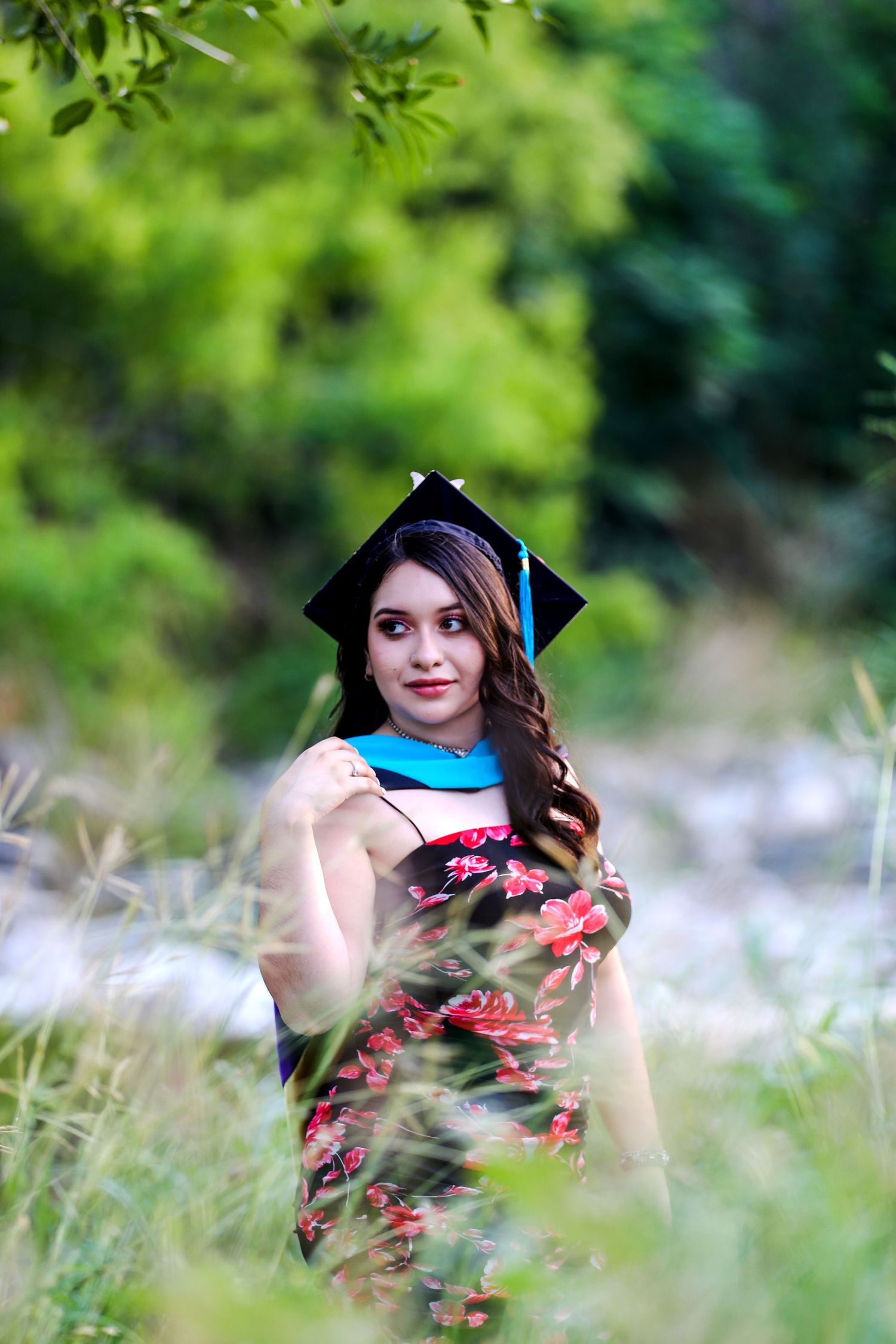 A graduation portrait taken outdoors with a black cap and blue sash amid blooming wildflowers and green foliage at Conception Park.