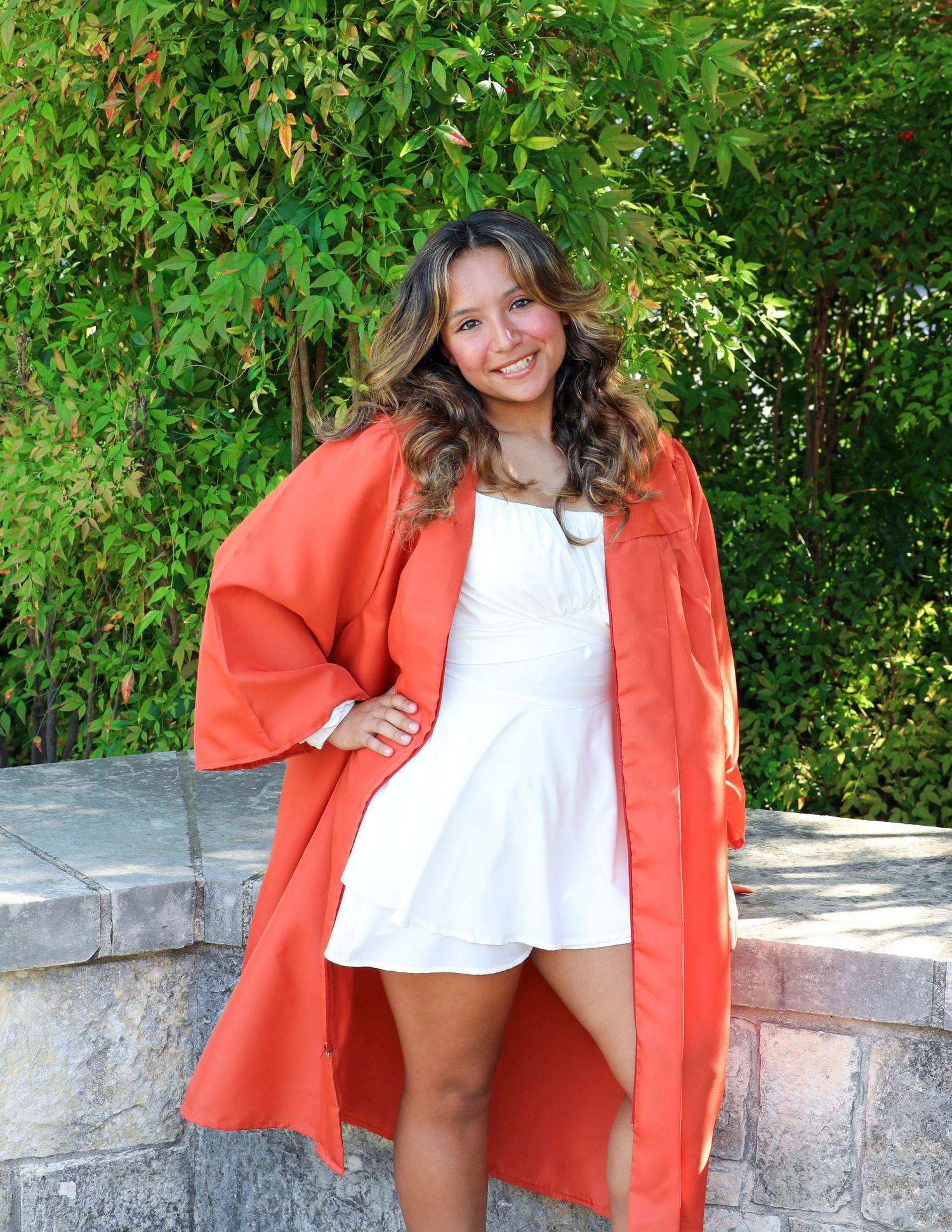 Smiling young woman in white dress and orange graduation gown posing outdoors near stone wall with green foliage.