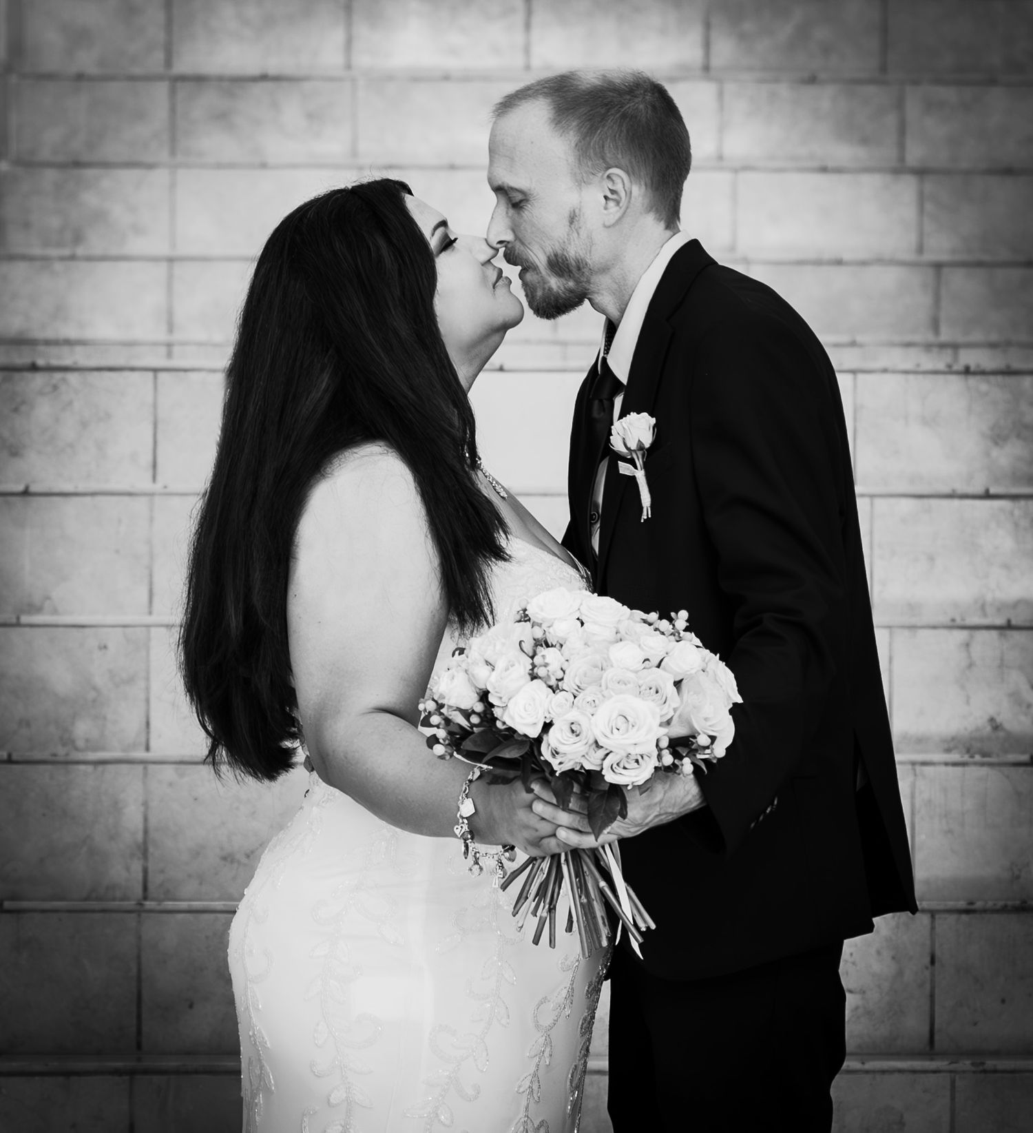 Black and white wedding photo of bride holding bouquet and groom sharing a kiss near a brick wall.