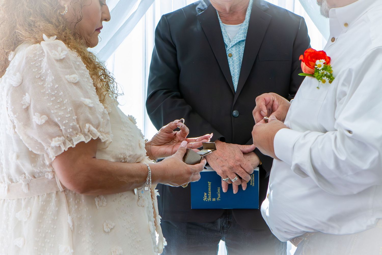 Couple exchanging wedding rings during an outdoor ceremony with an officiant holding a blue book.
