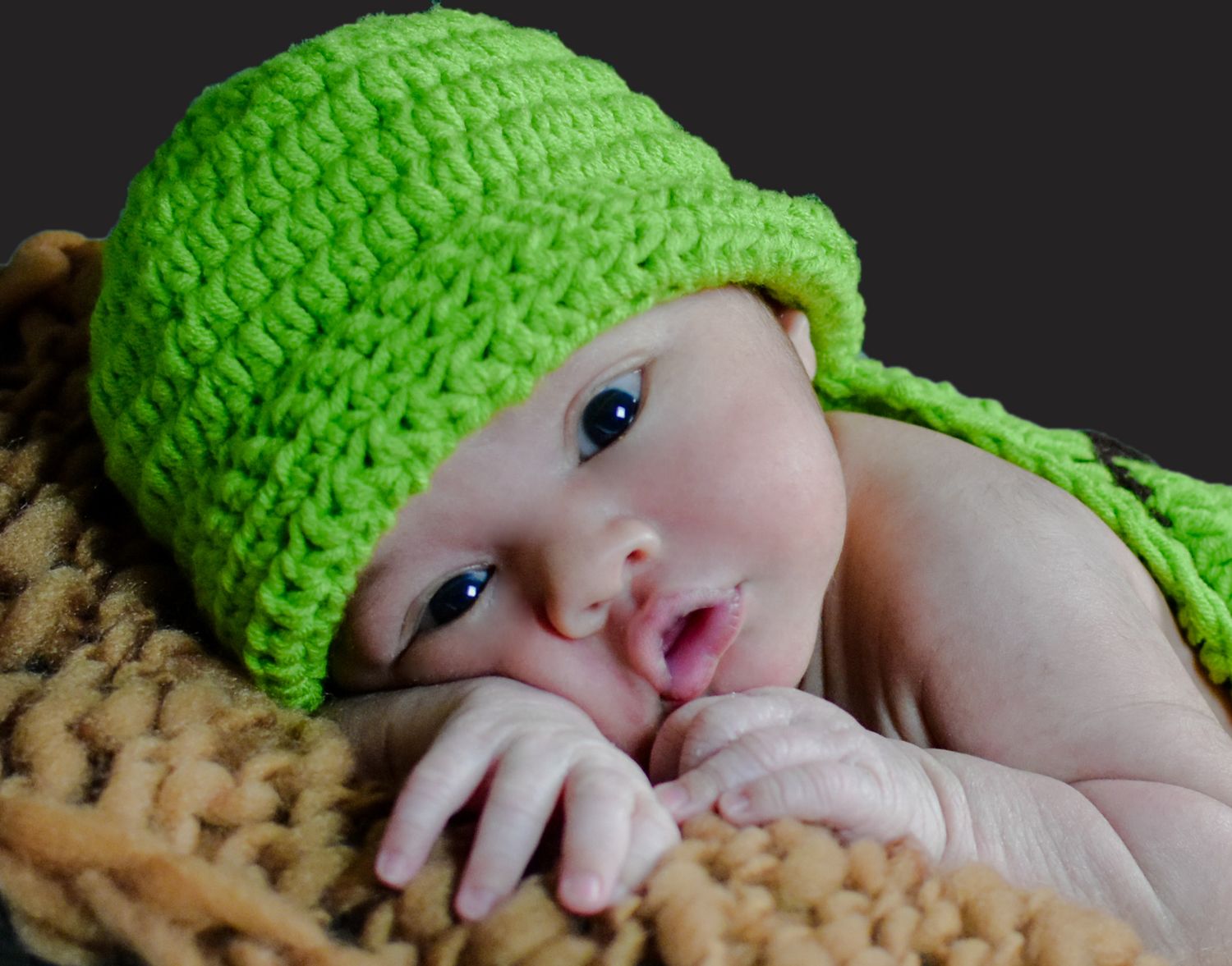 Adorable baby wearing a bright green crocheted hat lying on a soft brown blanket with wide curious eyes.