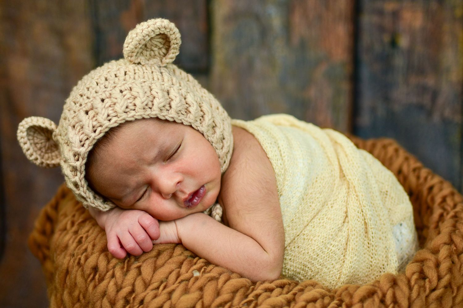 Sleeping newborn baby wearing a knit bear ear hat, nestled in a basket with a soft yellow wrap.
