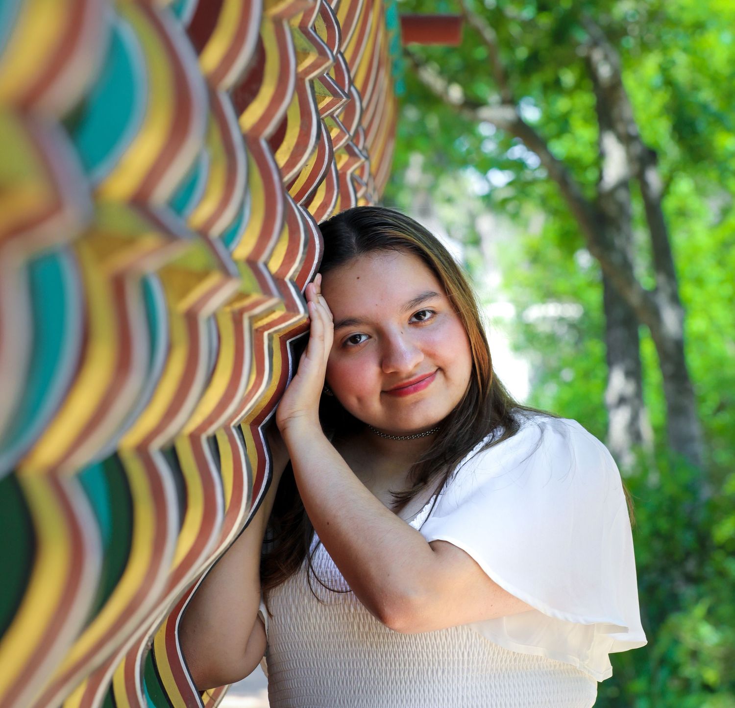 Young woman smiling beside a colorful striped art installation outdoors with green trees in the background.