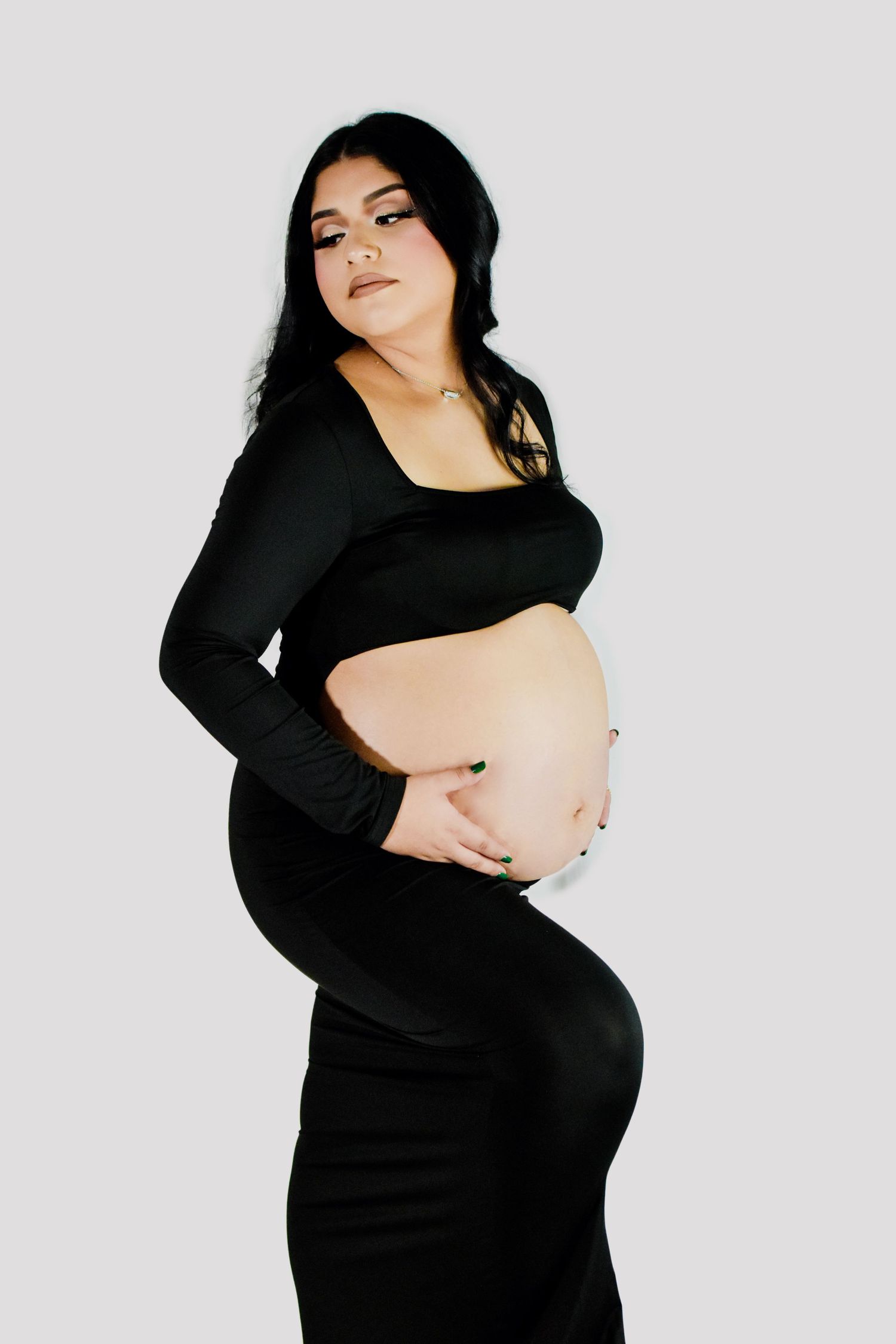 Pregnant woman in black crop top and skirt cradling baby bump, studio maternity photo on white background.
