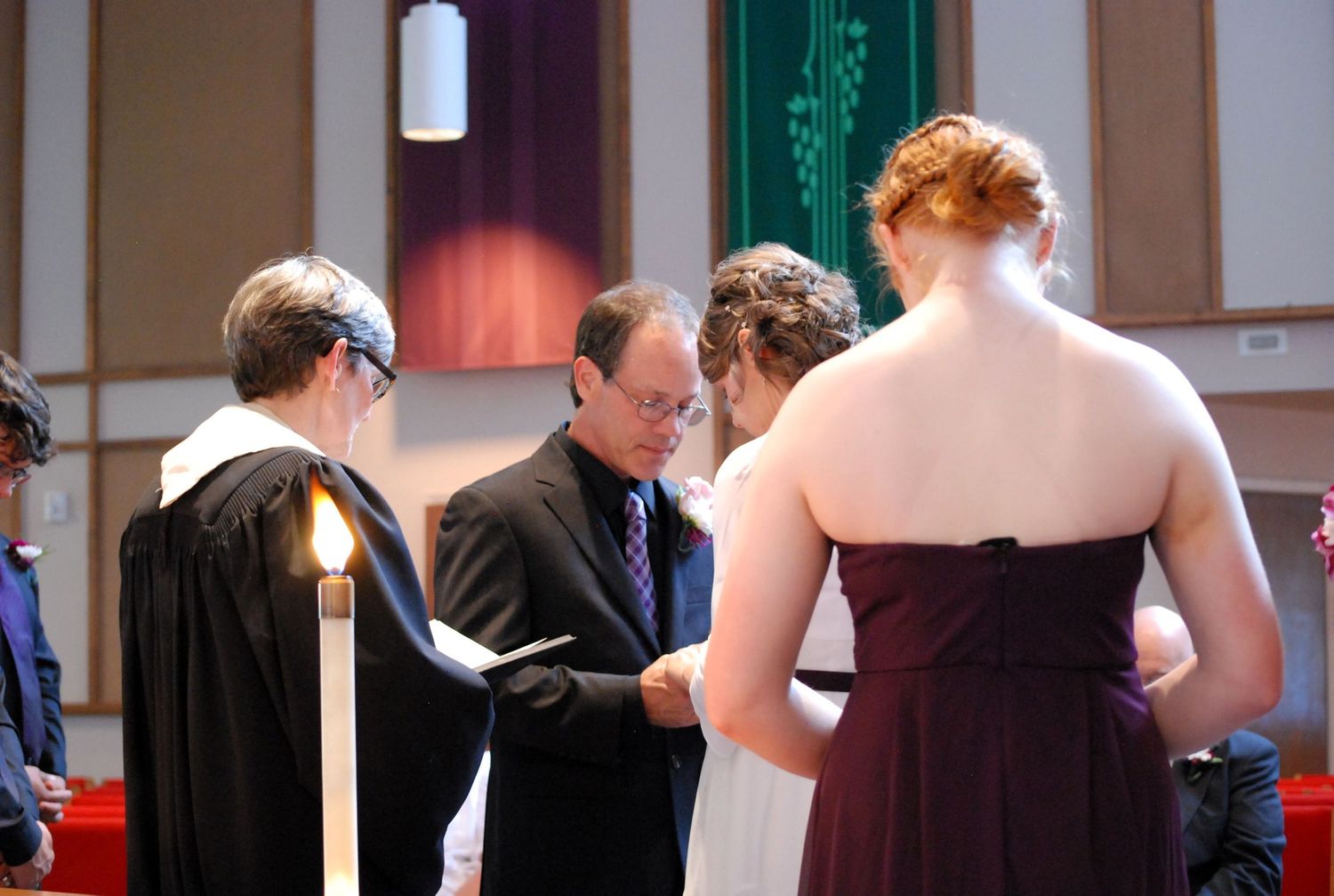 Wedding ceremony in a church with officiant, groom, bride in white, and bridesmaid in burgundy dress.