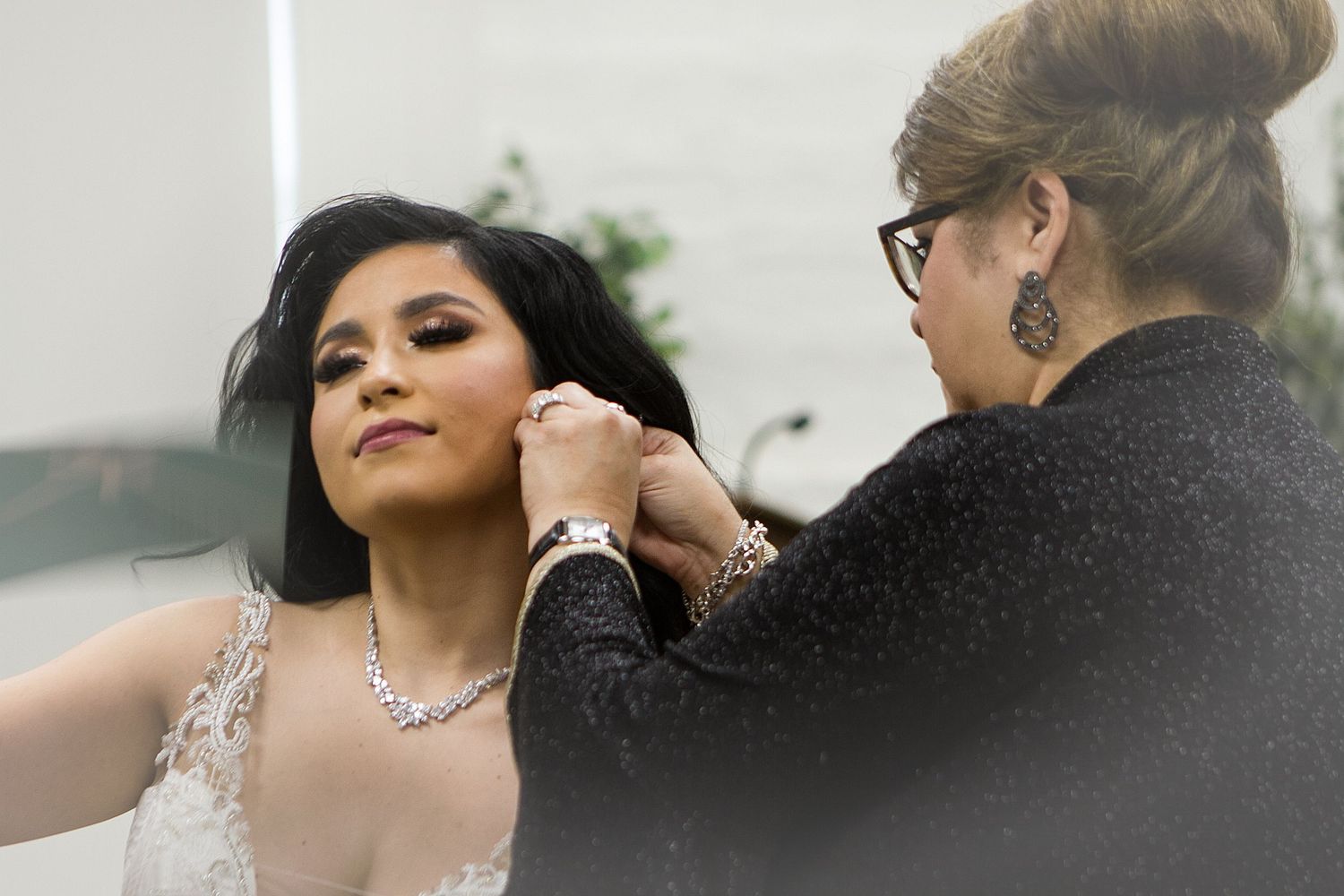 A woman in a wedding gown gets her necklace fastened by an older woman during bridal preparations.