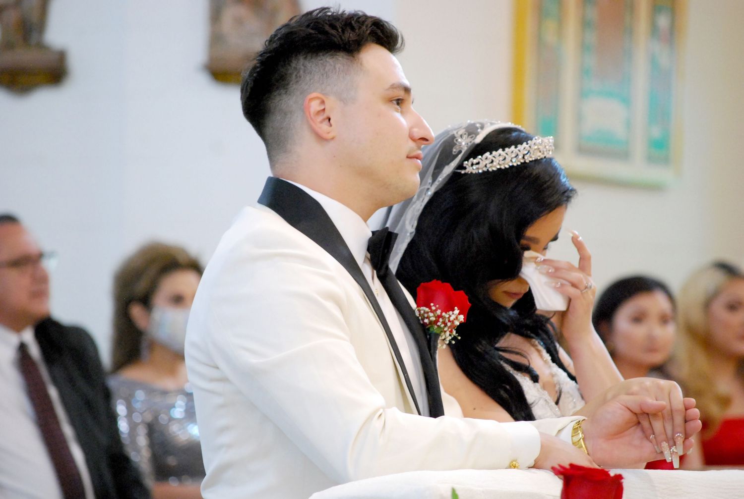 Bride wiping tears and groom in white tuxedo during emotional wedding ceremony at church altar.