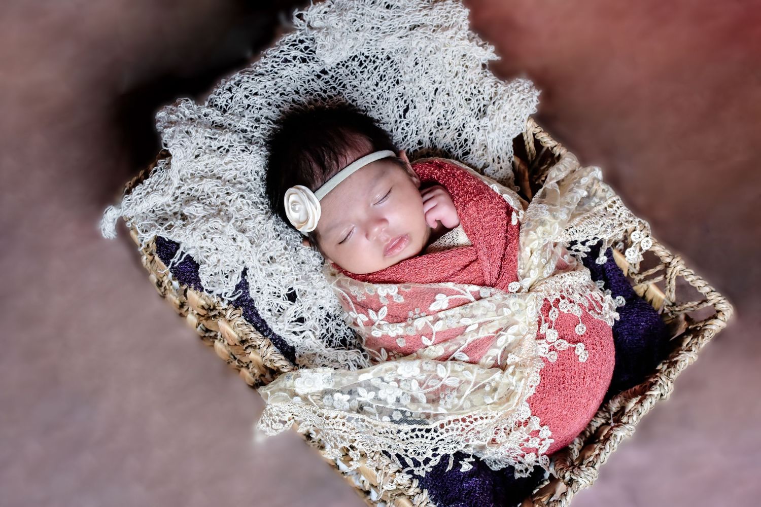 Sleeping newborn baby wrapped in pink lace swaddle with white bow headband resting in a rustic wicker basket.