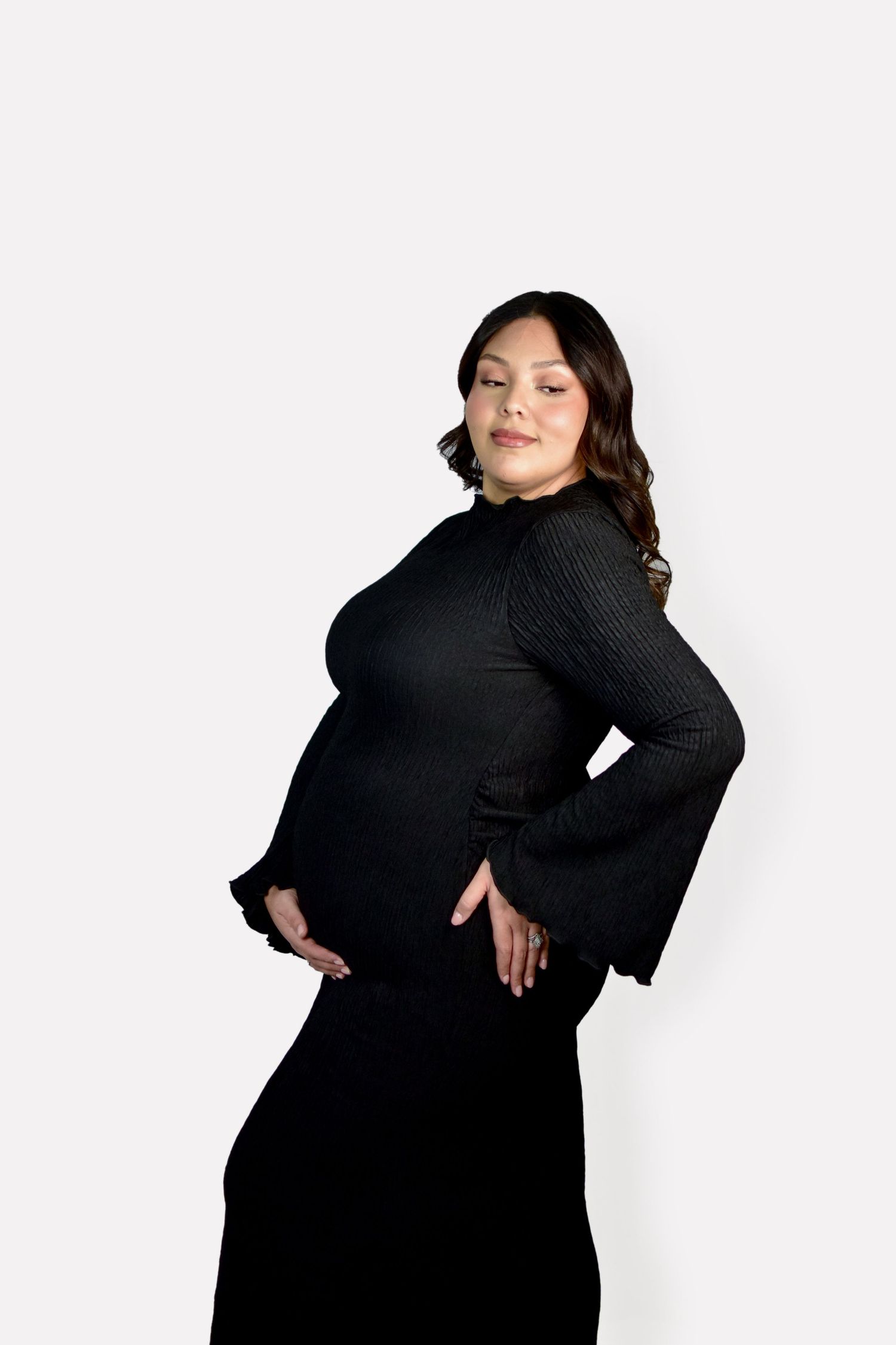 A mom to be wearing a black dress poses confidently against a white background at her maternity photo session in Austin, Texas