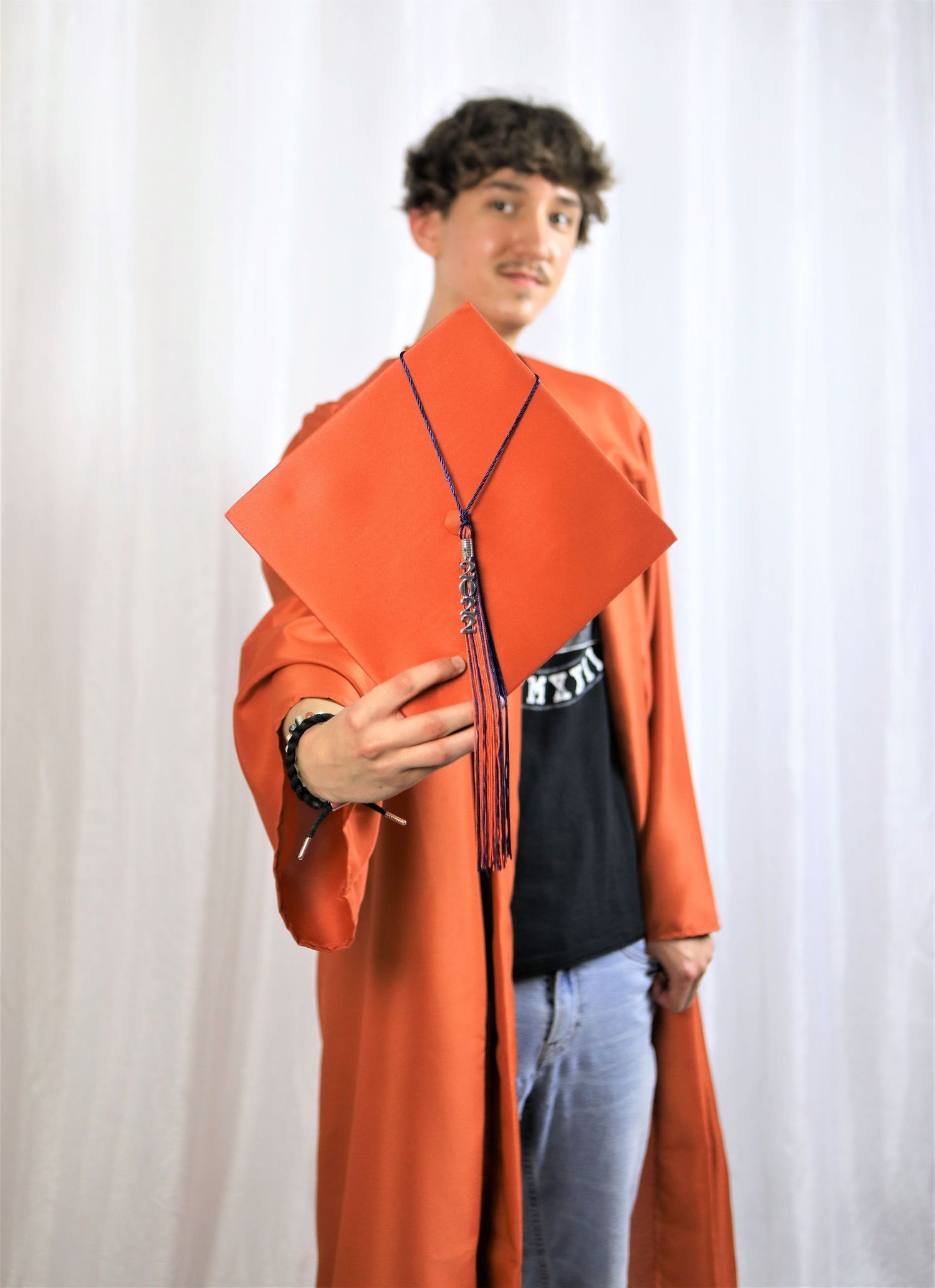 A student wearing a bright orange graduation gown holding his cap in studio setting for his senior photo session in San Antonio, Texas