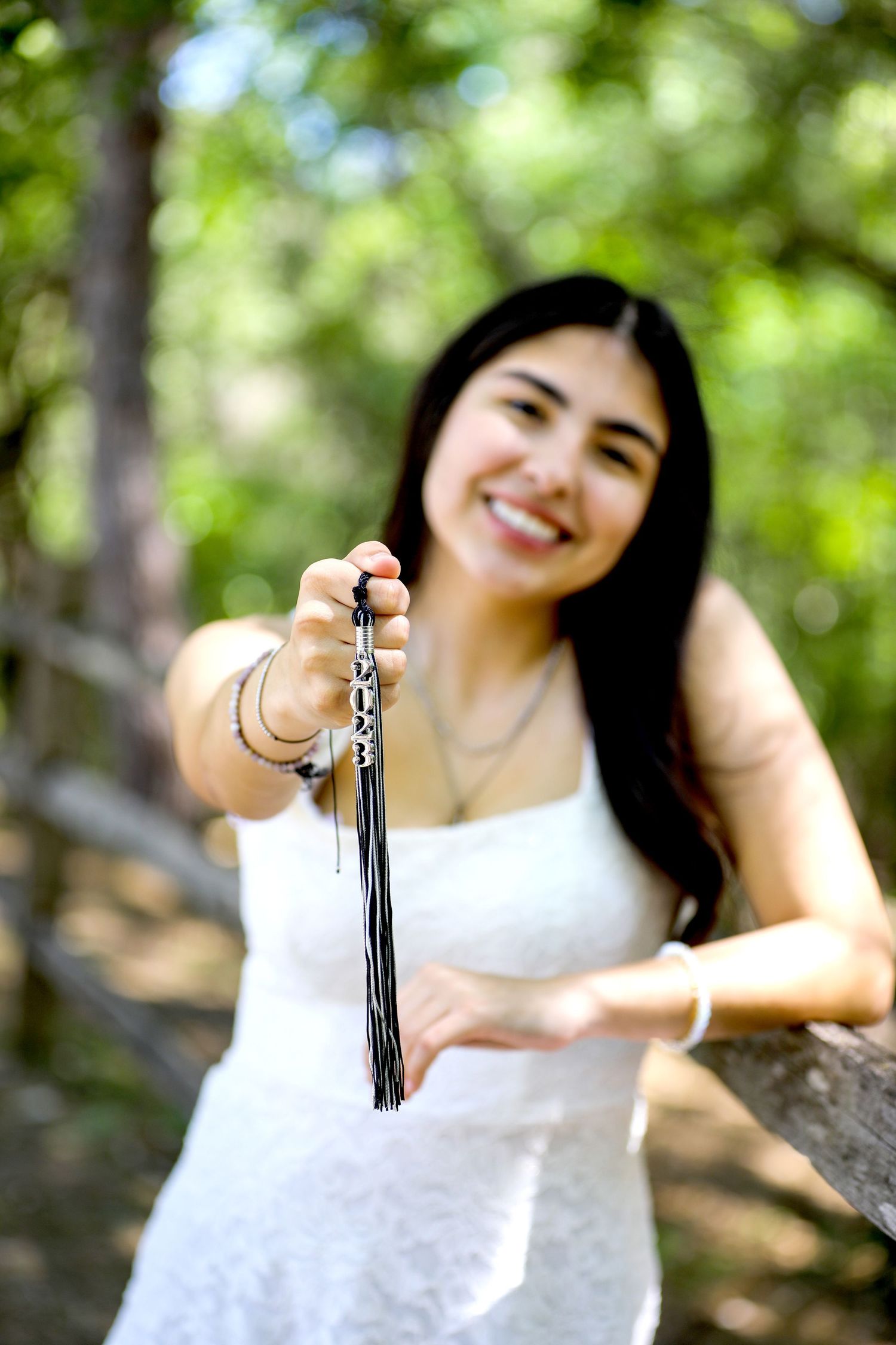 A student in a white dress poses playfully with tassel in a sunlit outdoor setting at Denman Park in San Antonio senior photography session