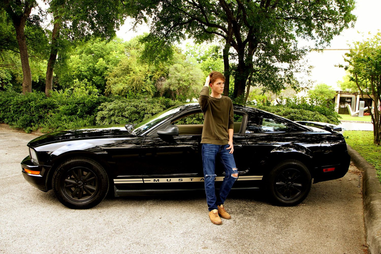 A student shows his graduation gift, a black sports car parked under green trees at the McNay Museum at his senior photography session.