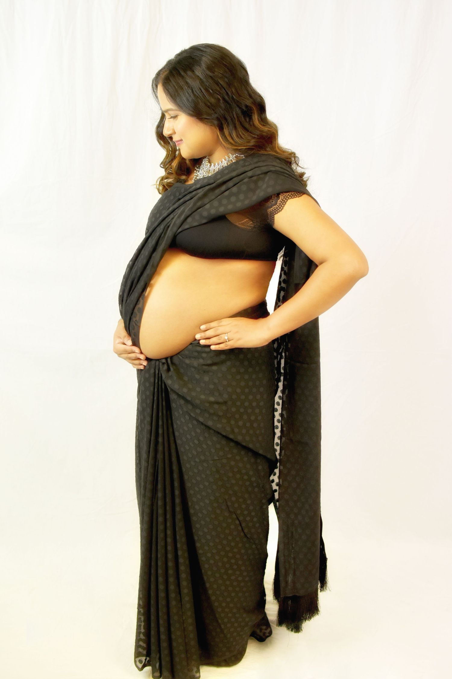 Pregnant woman in black saree cradling baby bump, posing gracefully against white background.